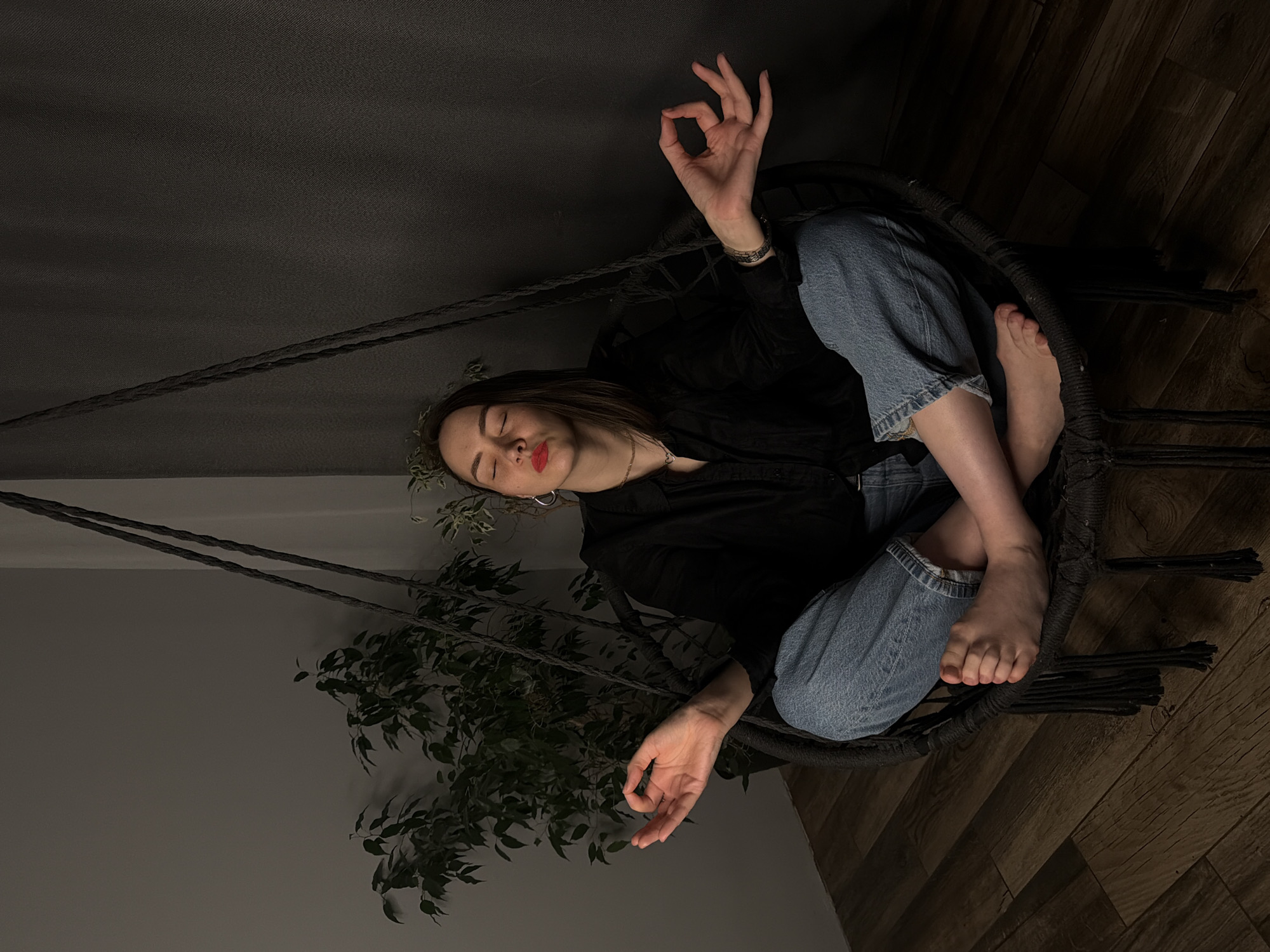A woman sits cross-legged in a hanging chair against a wall, indoors. She wears a black shirt and light-wash blue jeans with cuffed hems. Her eyes are closed. One hand is raised, forming an "OK" gesture. She has dark hair and wears small hoop earrings. The chair has black rope supports. A potted plant sits to the left of the chair. The floor is dark wood. The city of Chaikovsky, Russia, is not visually discernable in the image.