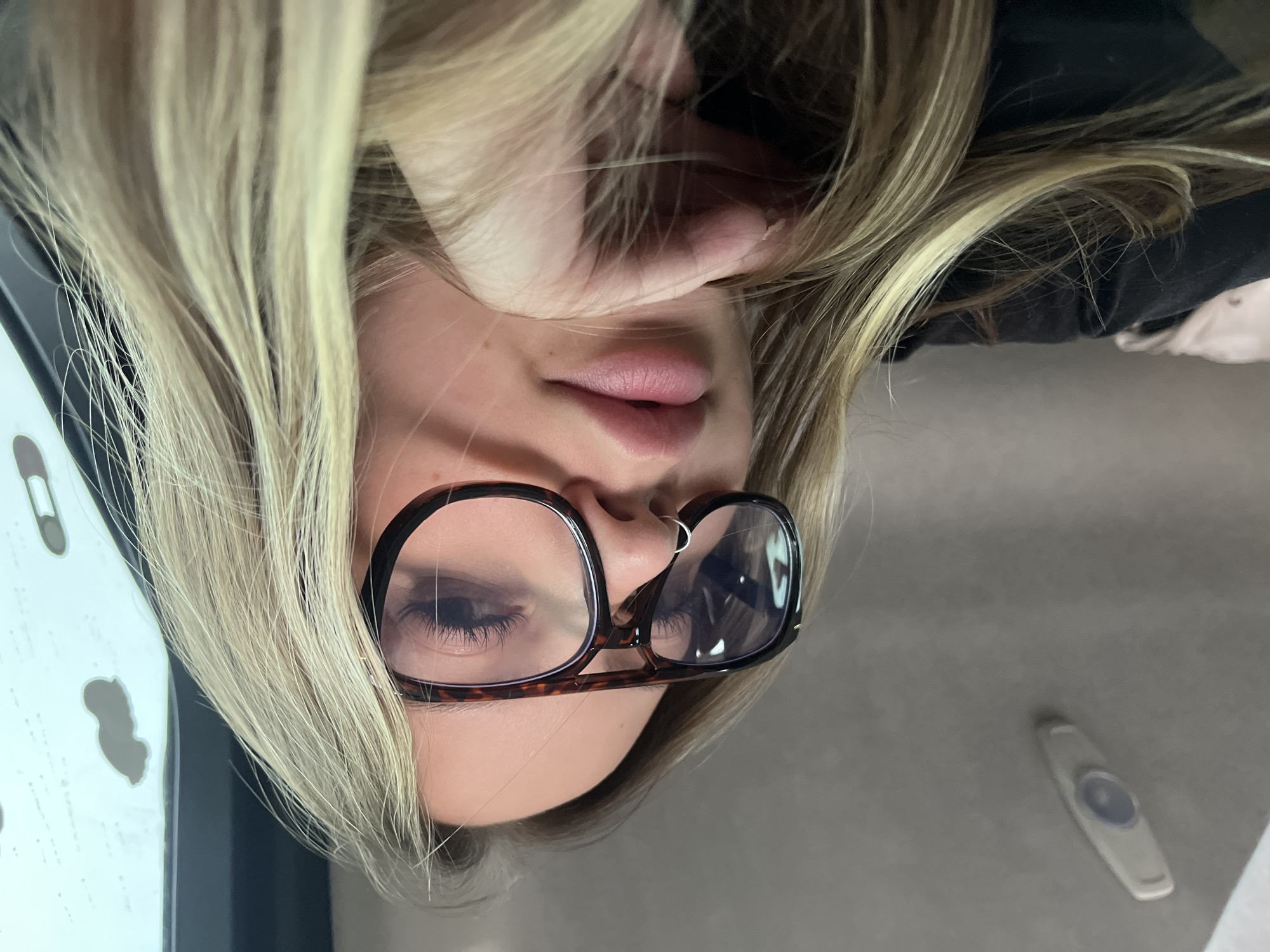 The image, which is rotated 90 degrees clockwise, features a close-up portrait of a person, likely female, inside what appears to be a vehicle in Staroderevyankovskaya, Russia. The individual has long, light blonde hair that cascades around their face and partially obscures their ear on the right side of the frame. They are wearing prominent tortoise-shell framed glasses, through which their eyes are visible, with some glare on the lenses. A small silver ring is piercing their left nostril, and their lips are slightly parted, indicating a neutral expression.
In the background, to the right of the person's head, a light-colored car headliner is visible, featuring an oval-shaped overhead light or control button. On the left side of the frame, there's a vertical light-colored surface with dark, irregular shapes, possibly a car interior panel or window frame. The person is wearing a dark-colored top, partially visible behind their hair. No text is discernible in the image. The scene captures a casual moment, possibly a selfie, within the car.