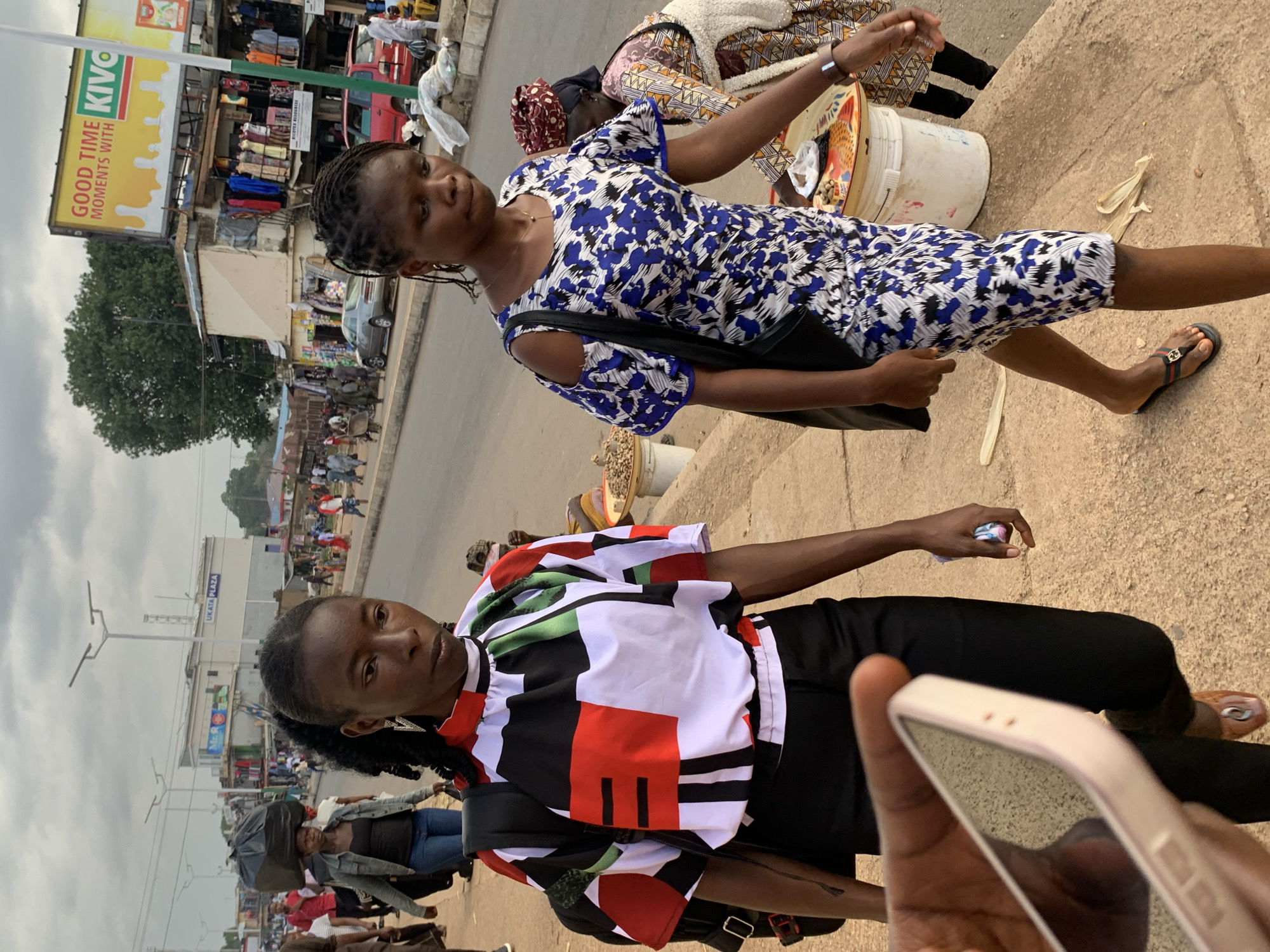 The media file depicts a street scene in Jos, Nigeria, with a focus on two young women. The image is oriented vertically, suggesting it might have been taken with a phone camera.
In the foreground, one woman is prominently featured, wearing a black garment with a colorful, abstract pattern of red, white, black, and green. She has braided hair and is looking towards the camera. A black bag with a strap is slung across her body. Her feet are clad in casual sandals. Her hand is visible in the bottom right corner, holding a light-colored smartphone, with the screen reflecting the surroundings.
Behind her, another woman is visible, wearing a blue and white floral print dress. She is also looking slightly away from the camera and appears to be walking or standing. She has a black bag over her shoulder.
The background reveals a busy street with various stalls and buildings, characteristic of a market area. Some of these stalls display clothing and other goods. There is a red vehicle in the mid-ground, and several other people are scattered throughout the scene, some carrying items. The overall atmosphere suggests a daytime setting, possibly overcast given the lighting.
Visible text includes "GOOD TIME MOMENTS WITH KIVO" on a large yellow billboard in the background. Another sign reads "UKATA PLAZA". The environment appears to be an urban street with shops and pedestrian activity. The women's expressions are neutral.