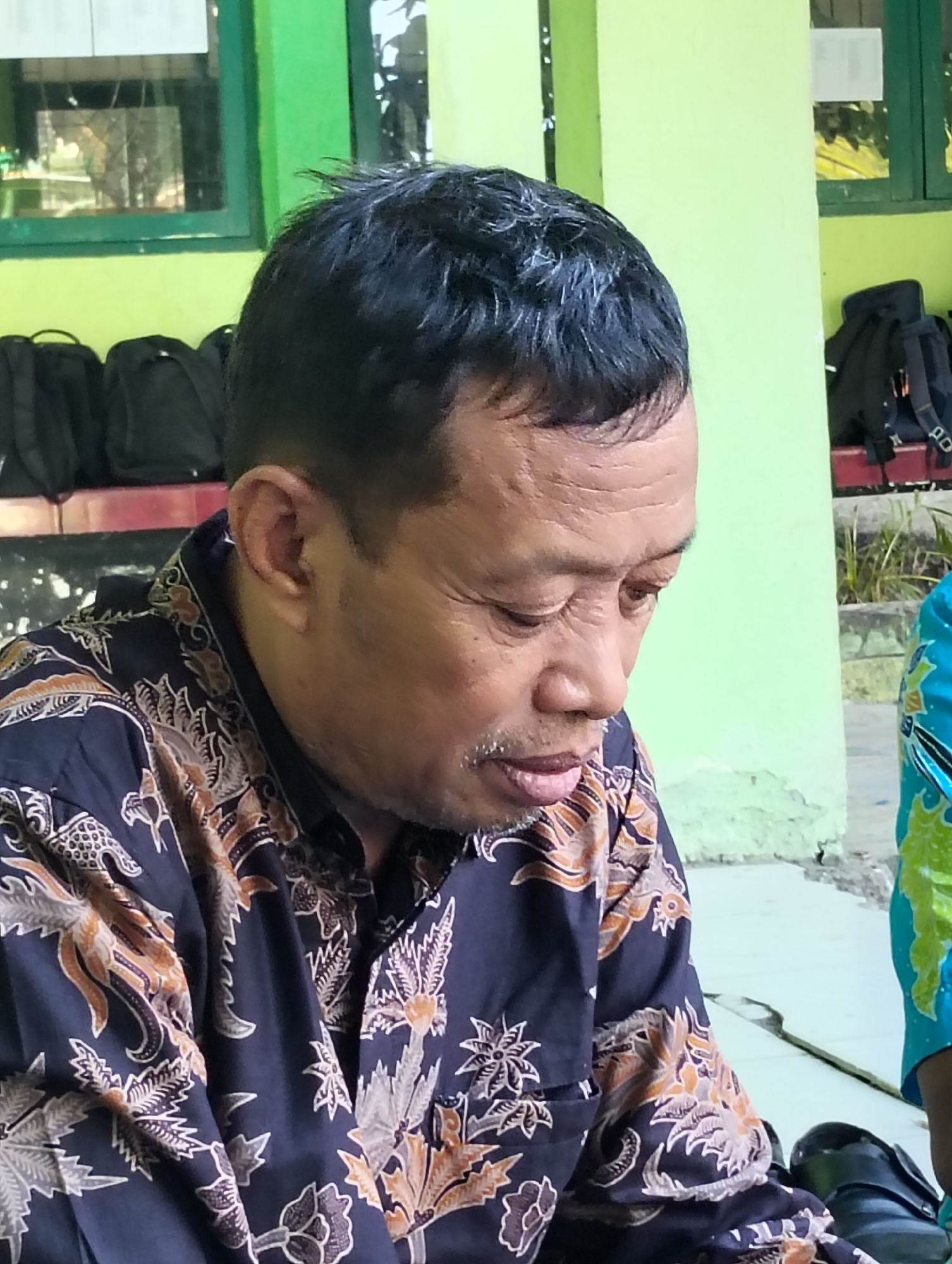 A man with short, dark hair and light brown skin is shown from the chest up, looking down and to the right. He is wearing a dark blue batik shirt with a pattern of orange and white stylized floral and avian motifs. He has a light mustache and goatee. The background is a light green wall with some peeling paint. To the left of the man, several black backpacks are visible, lined up against the wall. To the right, a portion of another person wearing a bright blue and green shirt is visible. The scene appears to be outdoors, possibly near a building with green window frames visible in the upper left background. This scene is located in Dagan, Indonesia.