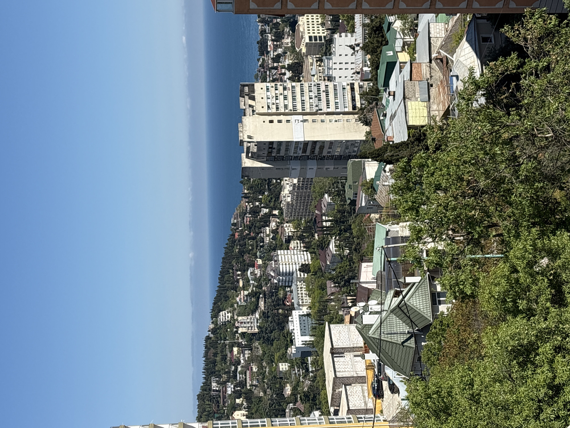 The image shows a view of город Ялта, Ukraine, from an elevated position. The sea is visible in the distance. The landscape slopes down towards the sea, covered in a mix of trees and buildings. There are residential buildings of varying heights and architectural styles. The image is taken from what appears to be a residential area. There are bushes and trees in the foreground. The sky is clear.