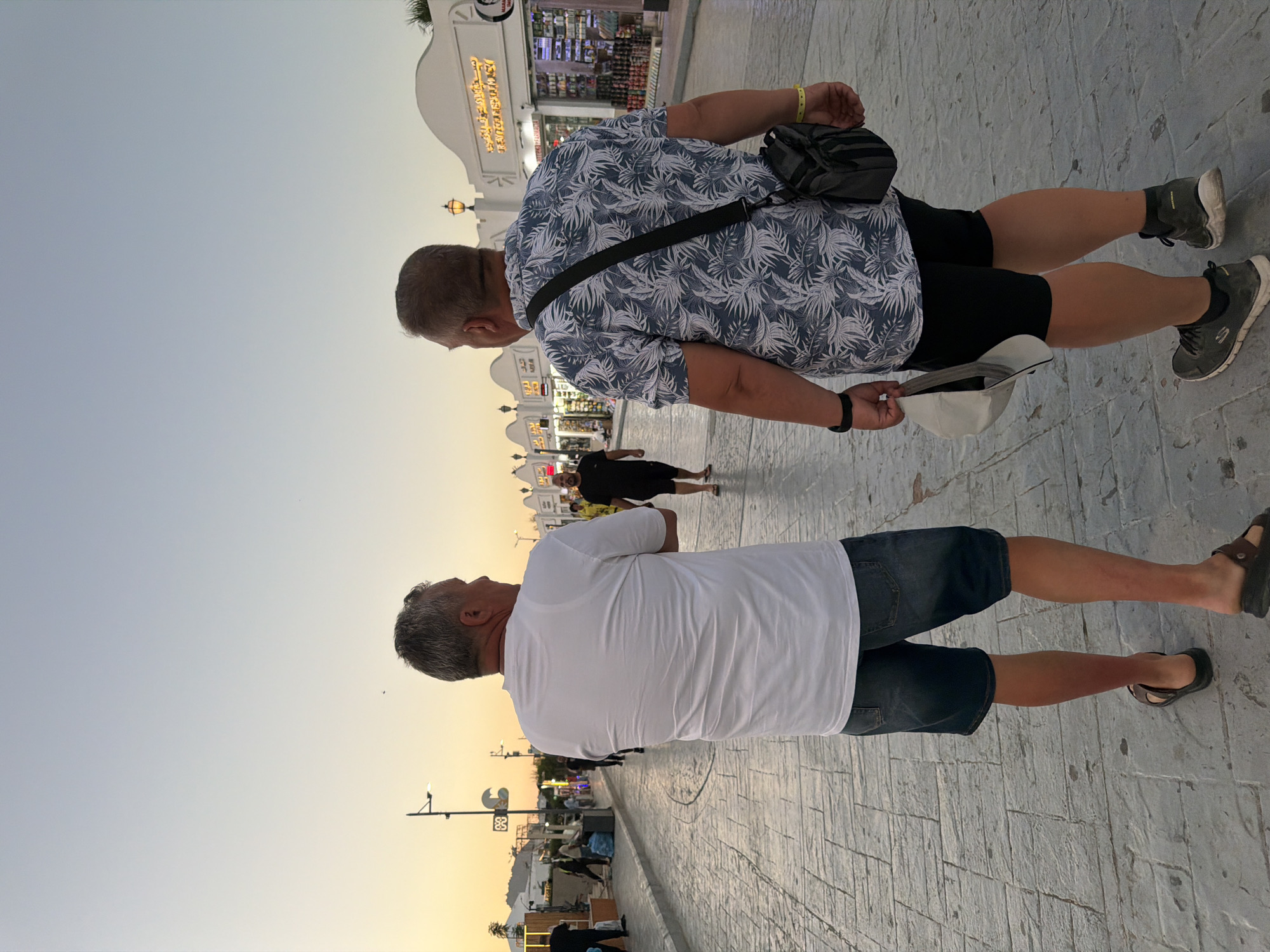 Two men are walking away from the camera on a paved street in Sharm El Sheikh, Egypt, at dusk. The man on the left wears a white t-shirt, denim shorts, and sandals. The man on the right wears a short-sleeved, patterned shirt, black shorts, and athletic shoes, carrying a cap and a small bag. The background features shops and streetlights, with a pale yellow and blue sky. Other pedestrians are visible in the distance, and the street is lined with buildings.