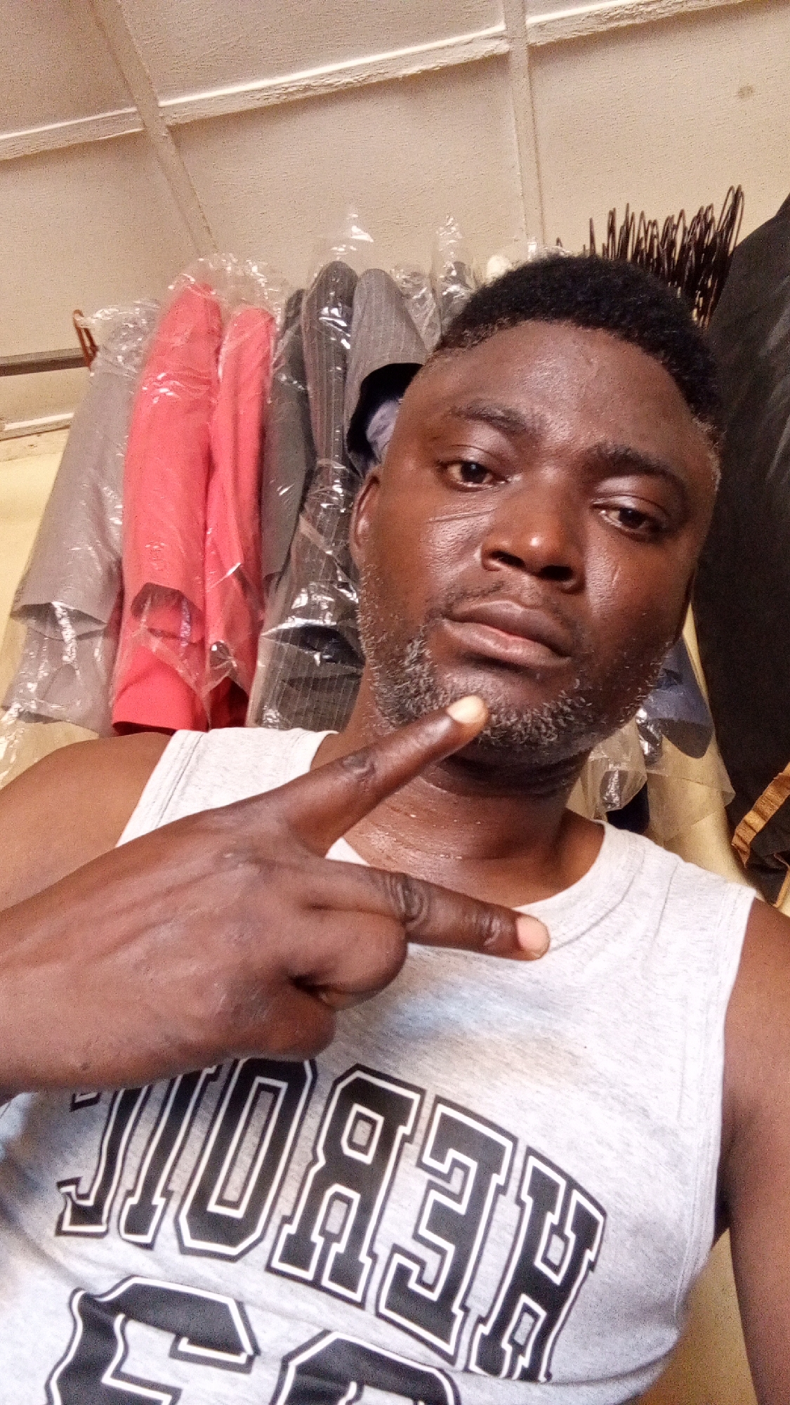 A man is posing for a selfie in what appears to be an indoor environment with clothing racks in the background. The man is wearing a grey tank top with a black and white graphic design on the front. He is making a hand gesture with his index finger pointing forward and his thumb pointing down. He has short, dark hair with some greying in his beard. The clothing racks behind him have various garments in plastic covers, with colors including red, grey, and striped patterns visible. The environment appears to be a shop or storage area. There is no definitive information to confirm this image was taken in Sabon Tasha, Nigeria.