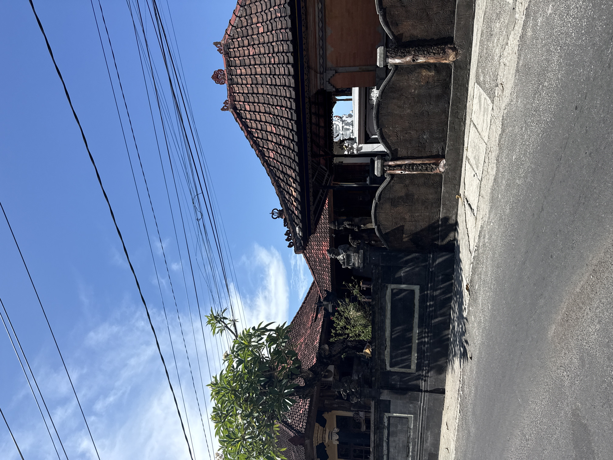 The image shows a street view in Batununggal, Indonesia, with traditional Balinese architecture dominating the scene. On the right, a section of a gray asphalt road is visible. Along the road, there is a building with dark stone walls, ornate wooden details, and a tiled roof with decorative elements. A tree with lush green foliage stands near the building. Overhead, a bright blue sky is crossed by several dark electrical wires.