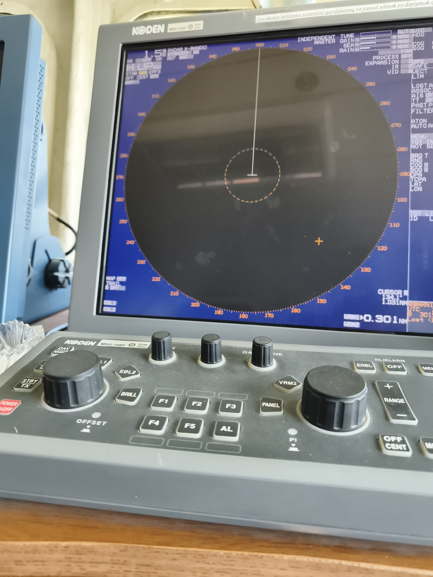 The image shows a close-up of a KODEN radar display and control panel, likely on a ship or a maritime vessel. The radar screen displays a circular sweep with various markings and numerical readouts, indicating a navigation or surveillance system. The control panel features several knobs, buttons, and labels, including "KODEN", "MRD-108P", "DAY", "NIGHT", "STBY TX", "POWER OFF", "OFFSET", "BRIL", "F1", "F2", "F3", "F4", "F5", "AL", "PANEL", "VRM2", "EBL2", "GAINE", "ERBL", "OFF", "HL/ALARM", "RANGE", "OFF CENT", and "MENU". The setting appears to be indoors, with a wooden surface in the foreground. The presence of maritime equipment suggests a location related to a port, harbor, or a vessel docked or navigating near Pirallahi Raion, Azerbaijan, given the context provided. There are no people visible in the image.