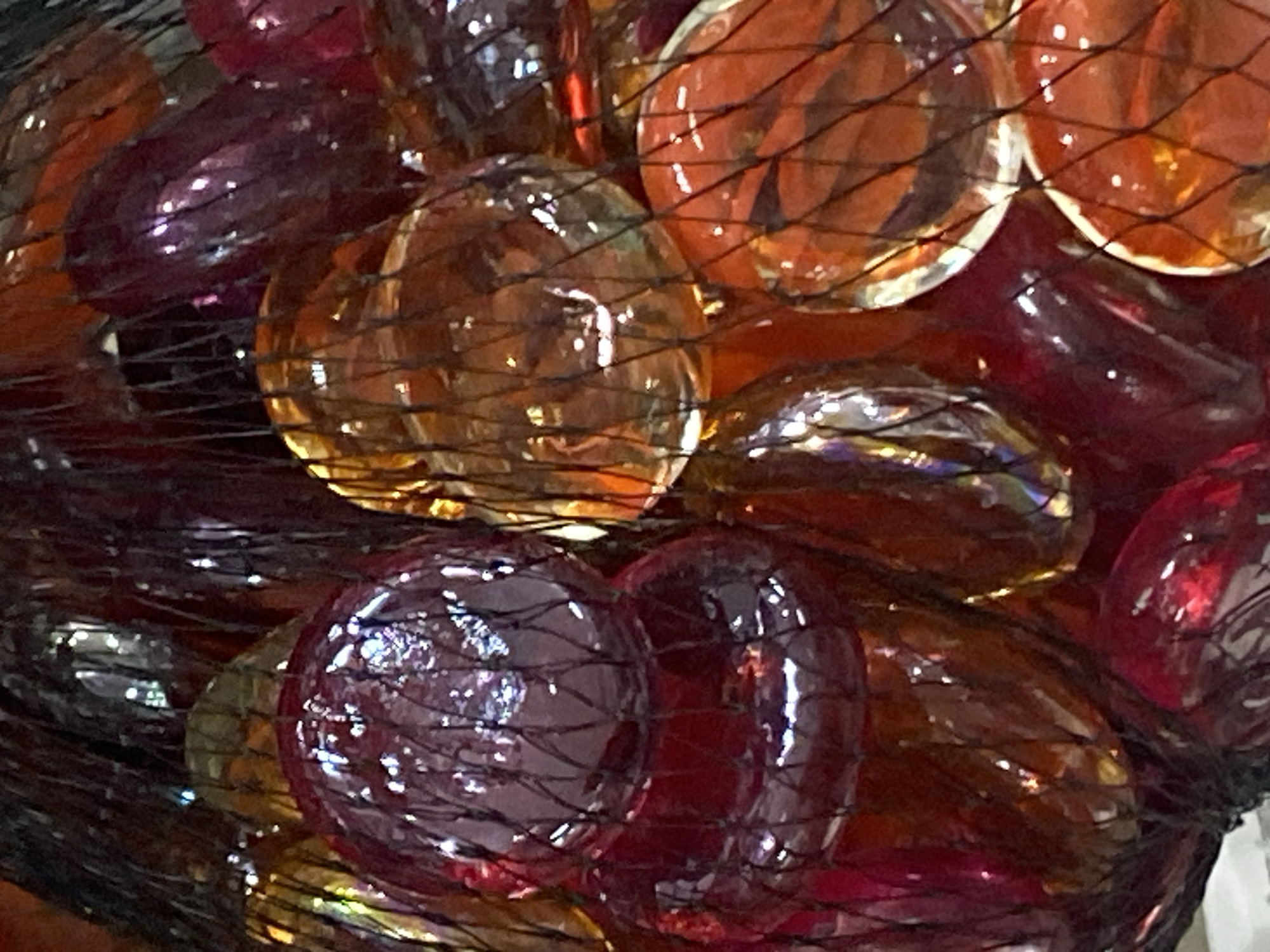 The image shows a close-up view of a collection of colorful glass beads, primarily in shades of red, orange, and purple, enclosed within a black mesh bag. The beads are round and appear to be smooth, with light reflecting off their surfaces, creating a shimmering effect. The mesh of the bag is visible, creating a grid-like pattern over the beads. The lighting is somewhat dim, contributing to the reflective quality of the glass. The scene appears to be an indoor setting, as suggested by the overall lighting and lack of natural environmental context. There is no indication of the location being in Krasnodar, Russia, or any specific city. The image is purely an object study, focusing on the texture, color, and light interaction of the glass beads and mesh.