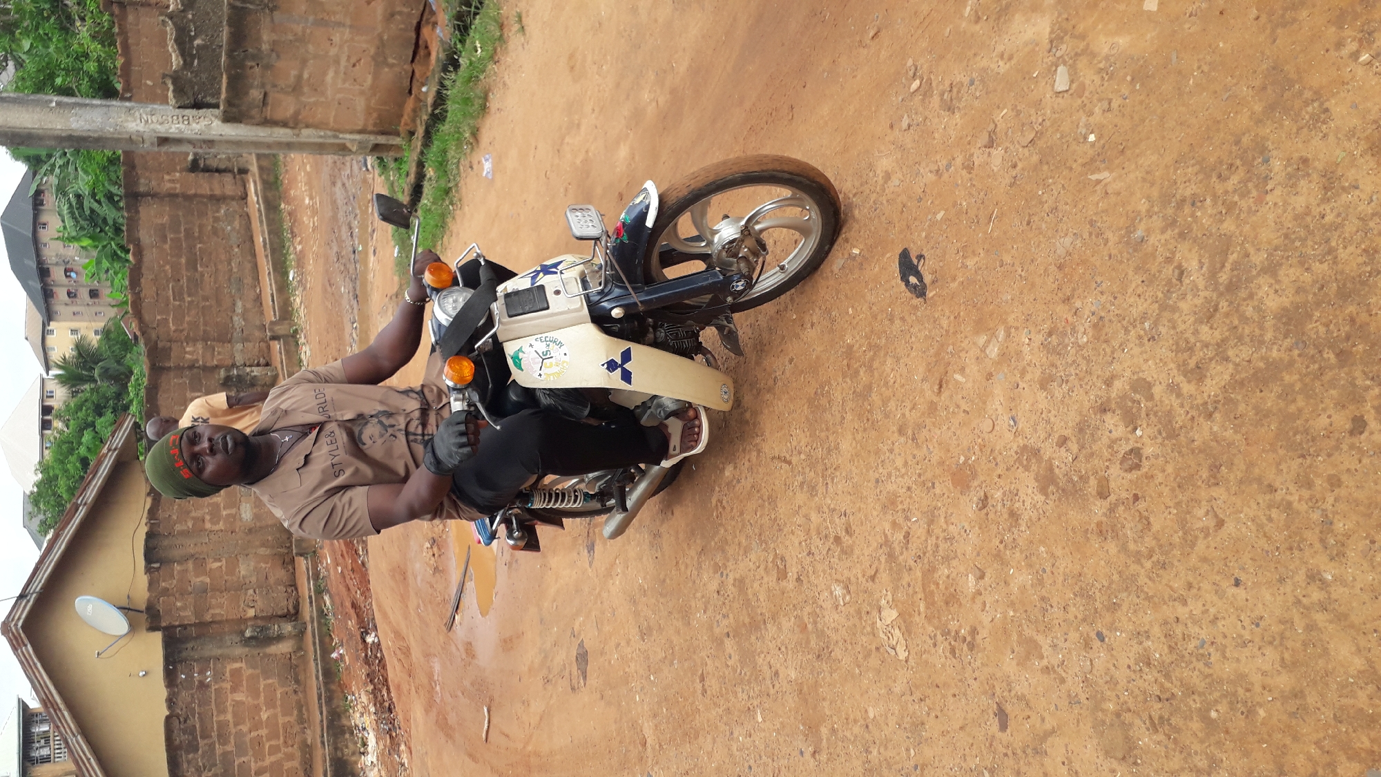 A man sits on a parked motorcycle in Nkpor, Nigeria. The ground is dirt-covered, and nearby buildings appear to be residential. The motorcycle is light blue and white, with distinctive decals. The man wears a brown shirt with black gloves and a dark hat. He sits looking toward the camera.