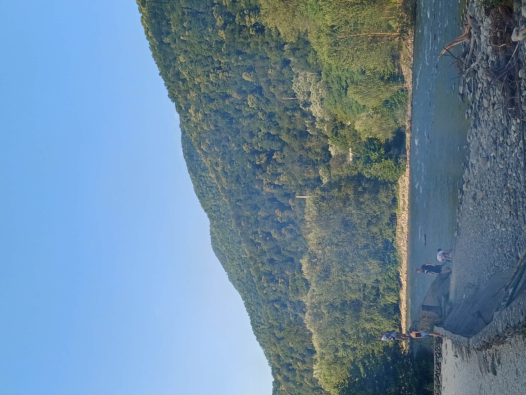 The image depicts a scenic river scene on a clear day, with a mountain covered in dense forest dominating the background. The sky is a vibrant, cloudless blue. In the foreground, a rocky riverbank lines a wide, flowing river. Several people are visible near the river. On the left side of the bank, four individuals are present. One person stands on a raised concrete structure, while three others are closer to the water's edge. Further to the right, two more individuals are near the water, with one of them appearing to be crouching down. The river itself shows ripples and movement, indicating a current. The overall environment is natural and suggests a recreational area by the water. The location is the city of Monastyr, Russia.