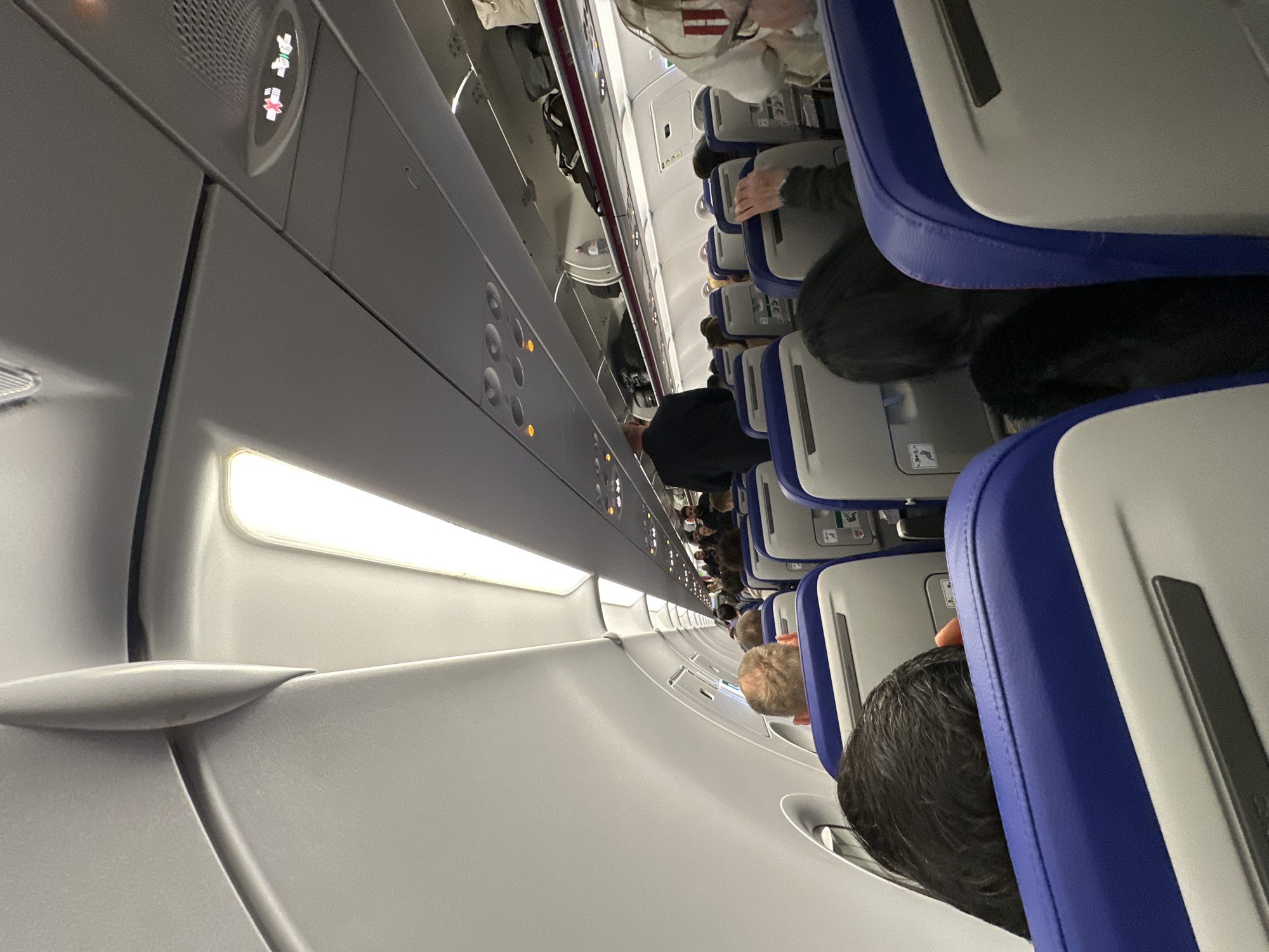 The image shows the interior of an airplane cabin. Passengers are seated in rows, facing forward. The seats are upholstered in blue and gray. Overhead compartments are visible along the ceiling. An aisle runs between the rows of seats. The lighting is bright. It is impossible to tell if it is from the city of Sofia, Bulgaria.