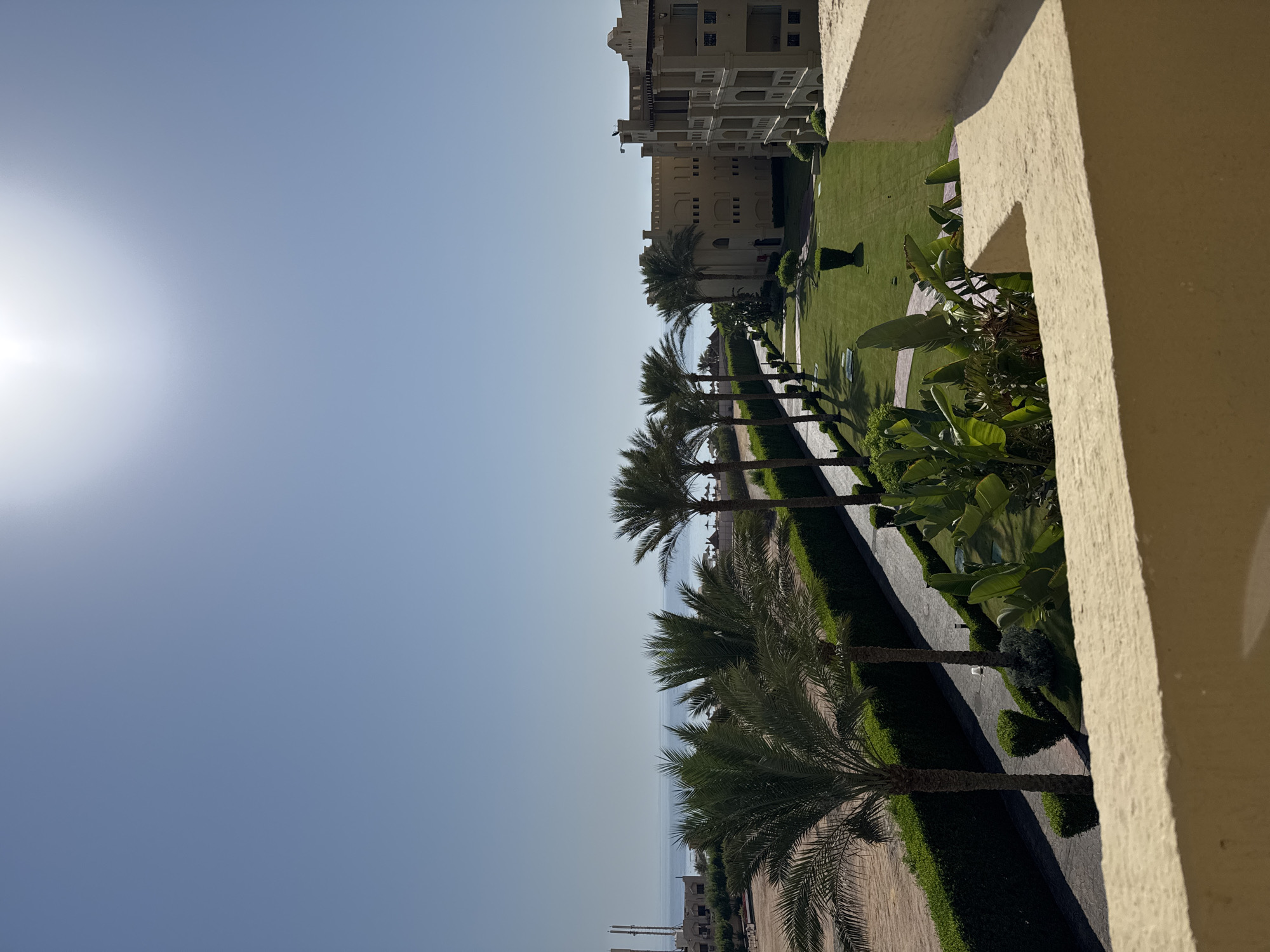 The image depicts a clear, sunny day with a bright sun in the upper right corner of a vast, light blue sky. In the distance, the calm, blue sea meets the horizon. Along the coastline, there are buildings and structures, including some that appear to be part of a resort or hotel complex, characterized by their light-colored, multi-story architecture and balconies.
In the foreground and mid-ground, a meticulously landscaped area features a manicured green lawn, a paved pathway winding through it, and several tall palm trees casting shadows. Dense green hedges line the pathway and the edges of the lawn. Lush tropical plants, with large green leaves, are also visible. The scene suggests a resort or hotel setting, likely in a warm climate given the presence of palm trees and the clear sky. The overall impression is one of a tranquil, well-maintained outdoor environment.