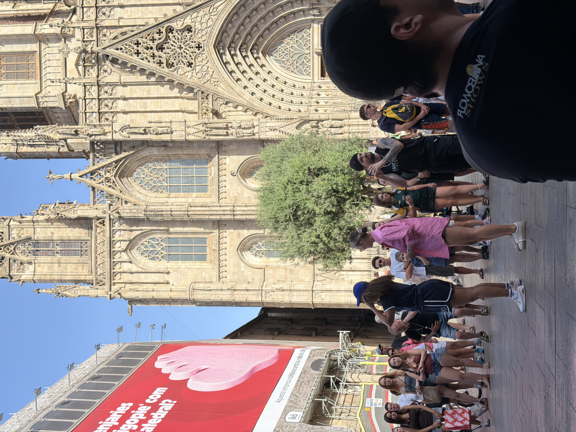 The image depicts a street scene in Barcelona, Spain, with the prominent Barcelona Cathedral in the background. A large red billboard with white text and a pink graphic of a hand is visible on the left side of the frame. In the foreground, a crowd of people is gathered on a paved plaza. Several individuals are standing and interacting, while others are walking. The architecture of the cathedral is a blend of Gothic and modern styles, with intricate stone carvings and stained-glass windows. The sky is clear and blue, indicating a sunny day. The overall atmosphere is lively and bustling, suggesting a tourist attraction or public gathering space.