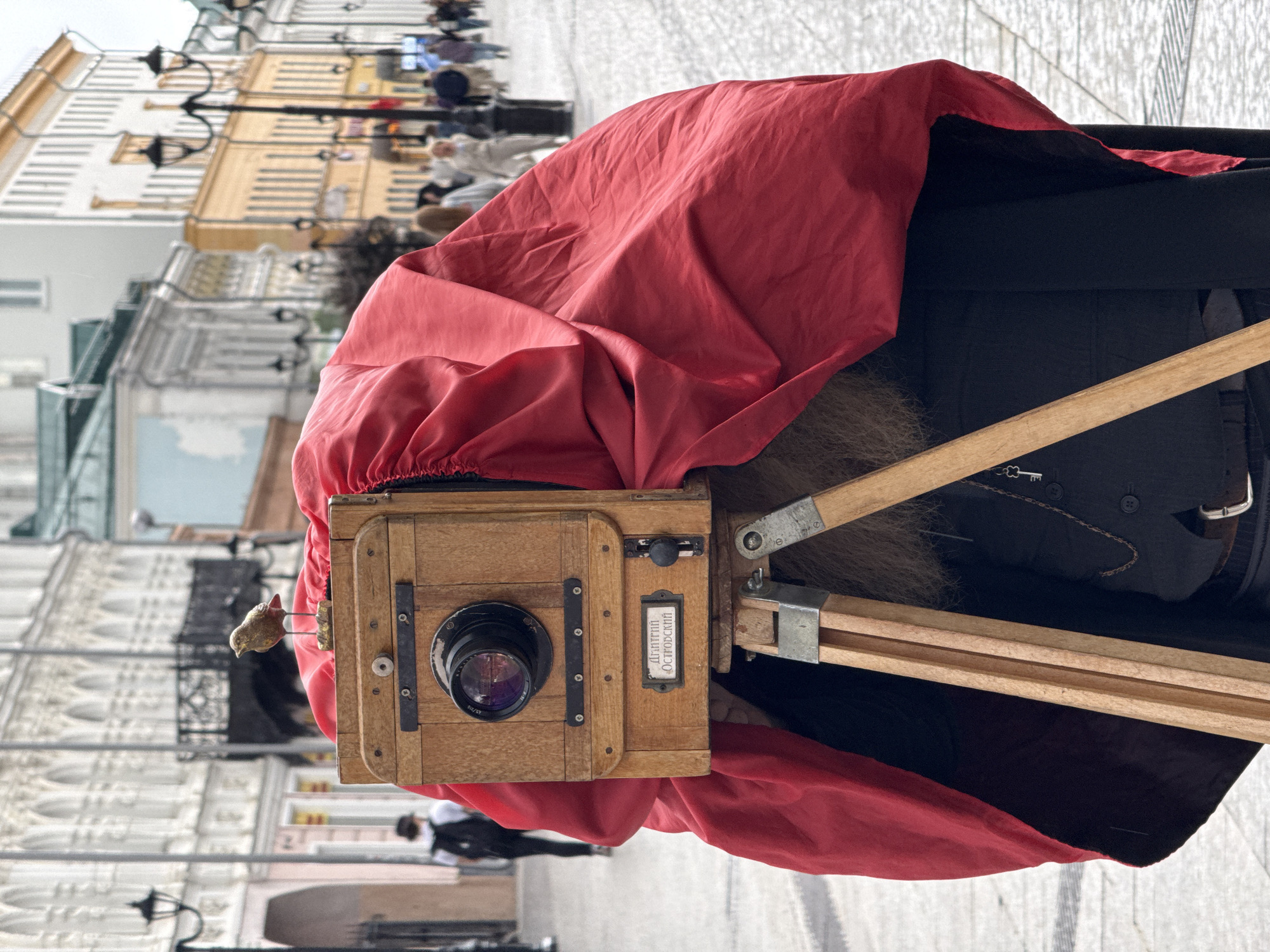 A street photographer, clad in dark clothing and covered by a large red cloth, operates a vintage wooden large-format camera on a tripod. A small, bird-shaped figurine is perched on top of the camera. The photographer's face is obscured by the cloth, but a hint of a beard is visible. The camera bears a label with Cyrillic inscription. The scene unfolds on a paved street lined with multi-story buildings featuring ornate architecture, characteristic of a historic city center. Street lamps are visible along the buildings. In the background, several blurred figures of pedestrians are seen walking. The overall atmosphere suggests a tourist or cultural district. This scene is located in Nizhny Novgorod, Russia, specifically on Bolshaya Pokrovskaya Street, known for its historic architecture and pedestrian activity.