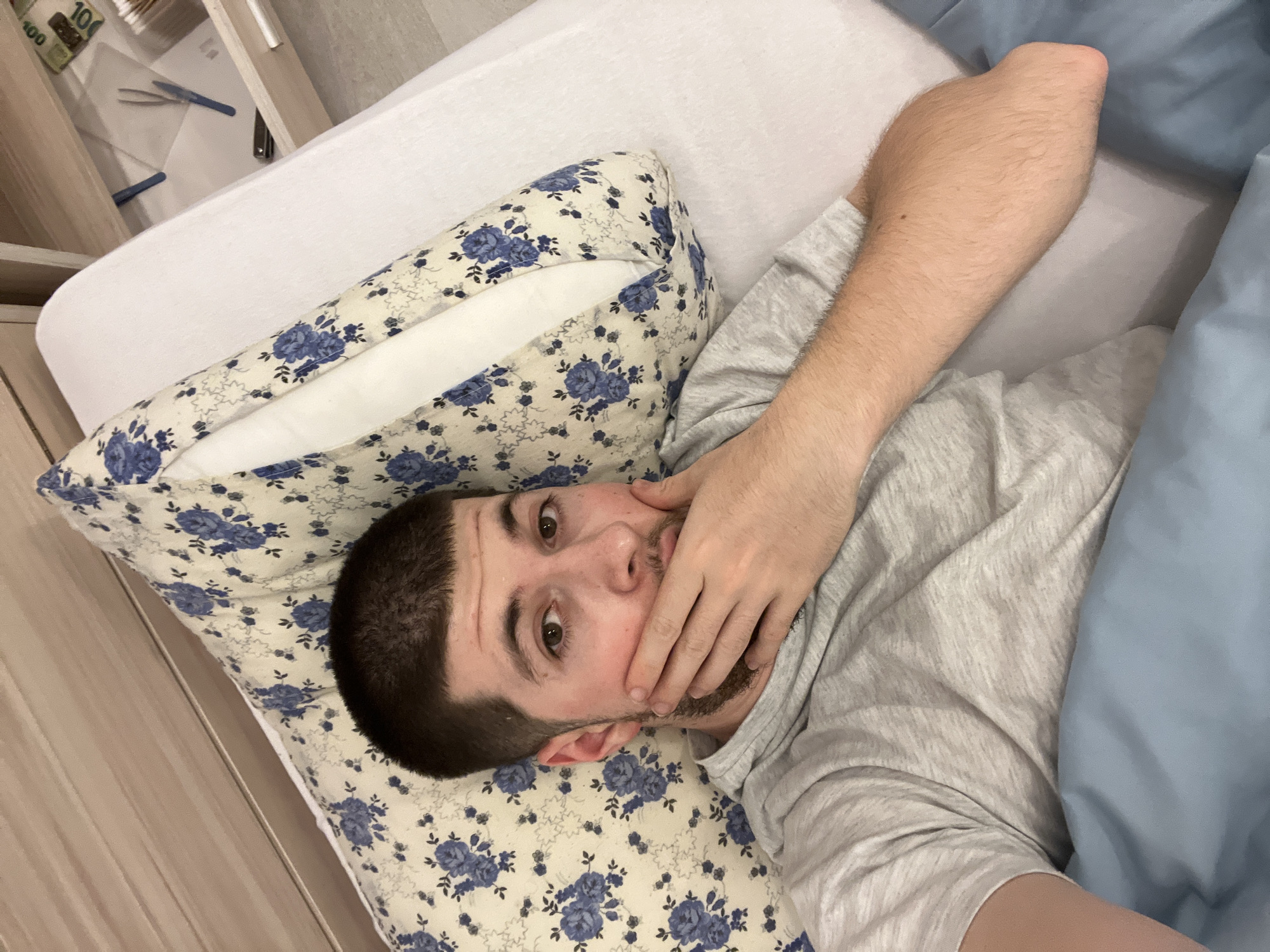 A man is lying in a bed. He is positioned on his back, with his head on a pillow with a blue floral pattern. His right hand is covering his mouth. The bed is covered with a white sheet and a blue blanket. The man is wearing a grey shirt. A bedside table is visible on the left side of the image, containing some items including paper, tweezers, pens, and money. The location is likely indoors, but there is not enough information to ascertain that the location is the city of Varna, Bulgaria.
