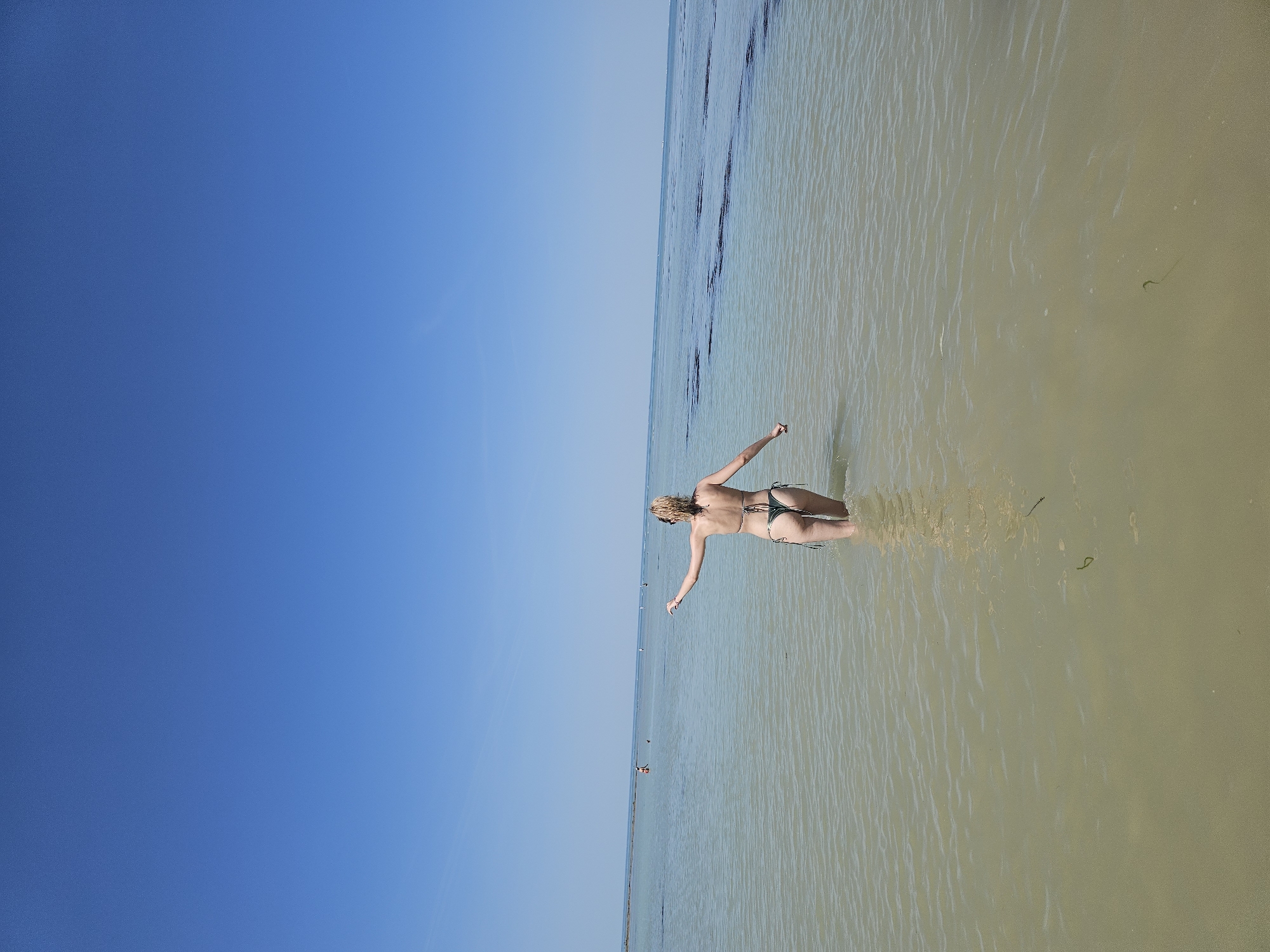 A vertical shot shows a woman standing in shallow water near La Brée-les-Bains, France. The water is light beige and murky. The woman, facing away from the camera, has her arms extended to either side. She is wearing a small dark bikini. The sky is a clear blue. The horizon appears to be straight and level.