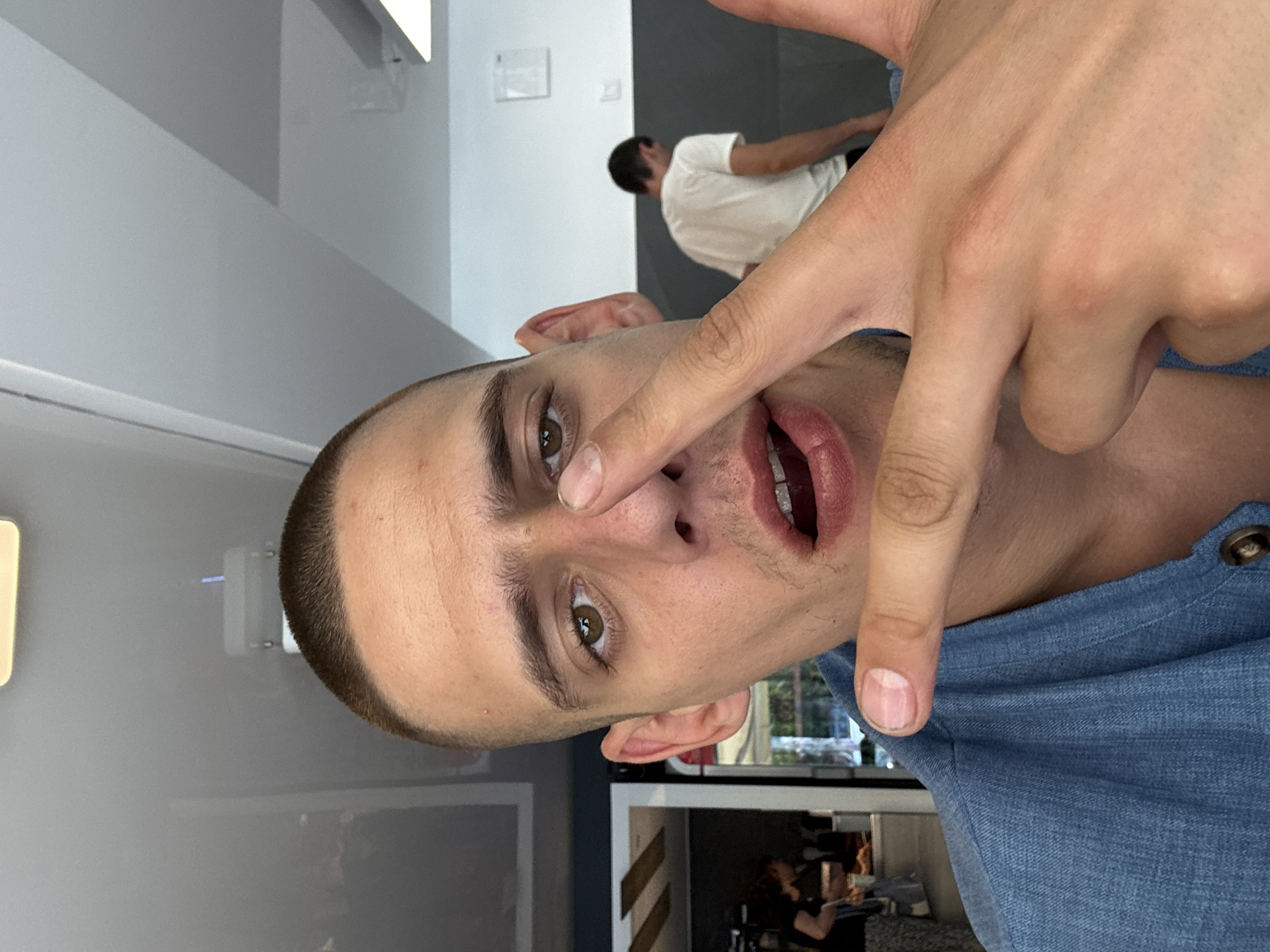 This is a close-up, first-person perspective photograph taken indoors, likely in a cafe or restaurant setting in Sofia, Bulgaria. The primary subject is a young man with a buzzed haircut and light brown eyes, looking directly at the camera. He is wearing a blue denim shirt. His right hand is raised, with his index finger touching his nose and his thumb and other fingers partially obscuring his face. His mouth is slightly open, revealing his teeth. In the background, there is a blurred image of another person wearing a white t-shirt, and further back, what appears to be a serving counter with other individuals present. The environment appears to be illuminated by overhead lighting.