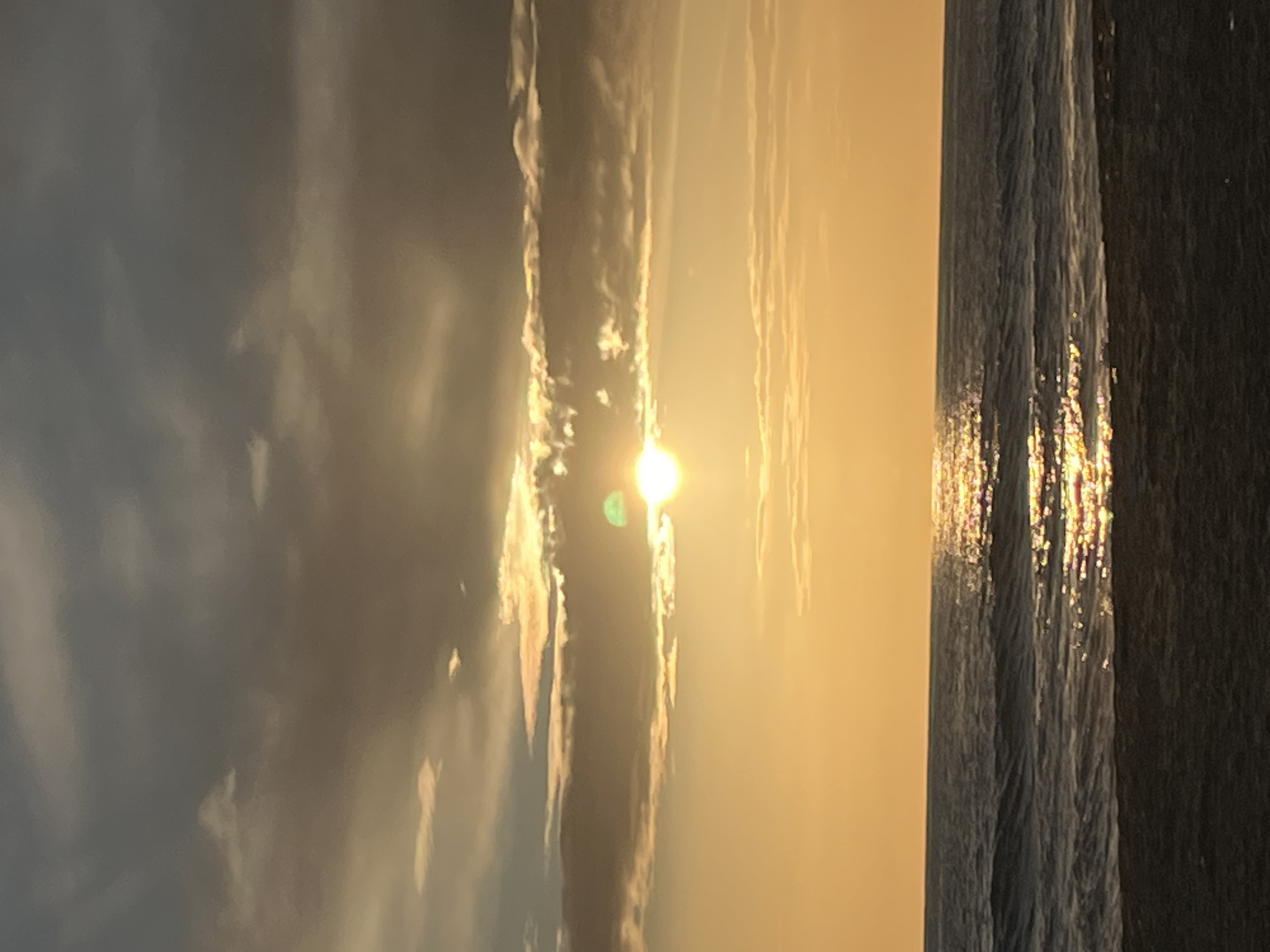 The image captures a vertical view of a beach scene, likely taken during sunrise or sunset in Alanya, Turkey. The sky is predominantly golden, with the sun shining brightly through clouds, casting a hazy glow. To the right, waves ripple across the water, reflecting the sunlight in a sparkling pattern. The sky appears to have varying shades of grey as well. No people are visible. The photograph emphasizes the natural beauty of the coastal environment.