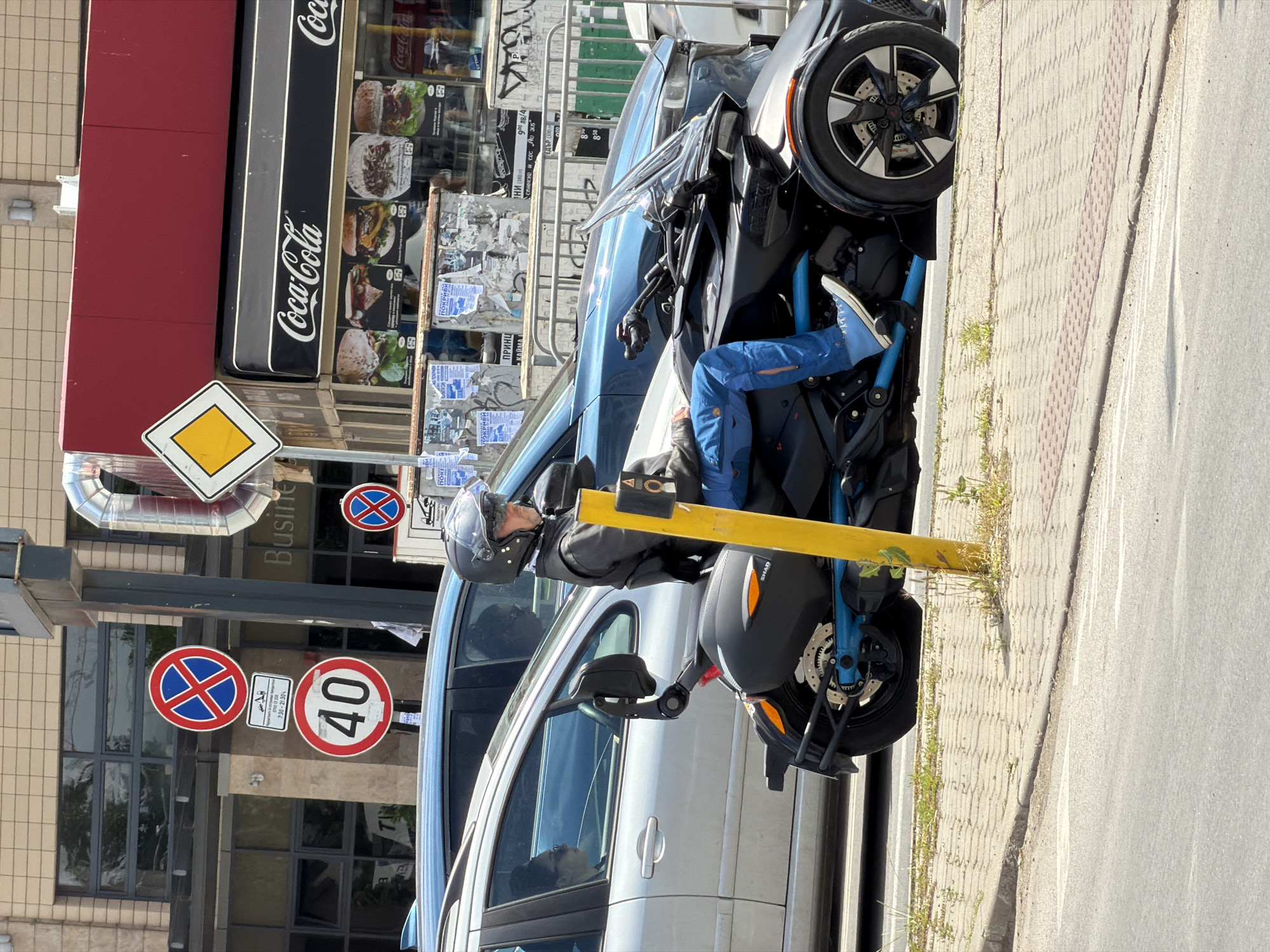 A man wearing a motorcycle helmet sits astride a black Can-Am Spyder three-wheeled motorcycle parked on a sidewalk in Sofia, Bulgaria. The motorcycle is parked adjacent to a silver van and a yellow bollard. The storefront behind the motorcycle features a Coca-Cola sign and a variety of food advertisements. Various traffic signs, including a "no stopping" sign and a speed limit sign (40 km/h), are visible along the street. Several posters and advertisements are plastered on the wall behind a metal fence. The overall scene appears to be an urban street environment.