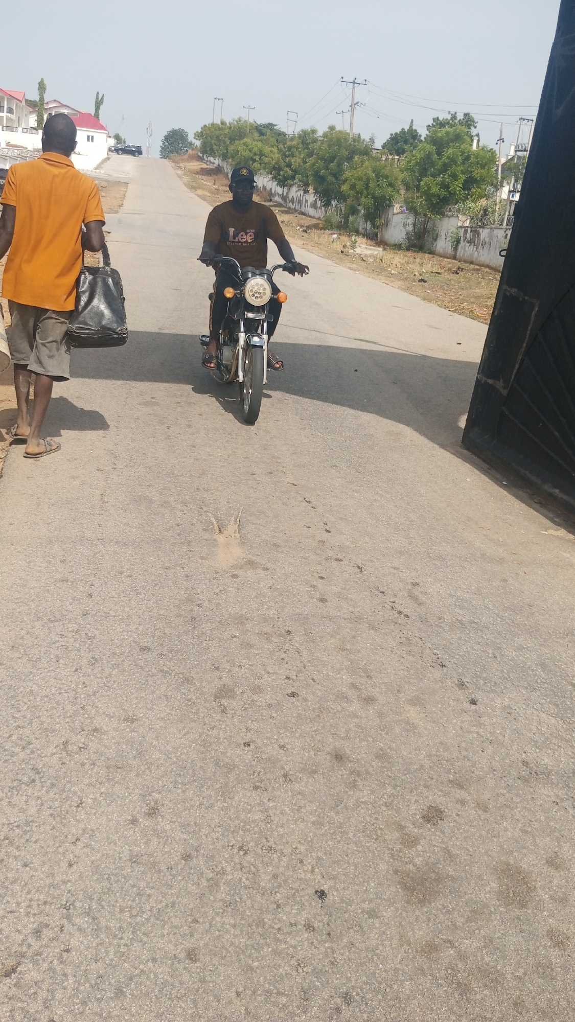 This image captures an outdoor street scene, likely in a semi-urban or residential area of Lokoja, Nigeria, under bright daylight conditions. The sky is clear and sunny, suggesting midday or early afternoon.
Two men are visible in the frame. On the left, a man is walking away from the camera, wearing an orange-brown t-shirt, grey shorts, and sandals. He carries a dark, possibly black, bag in his left hand or slung over his shoulder.
Further down the paved road and slightly to the right, a second man is riding a motorcycle towards the viewer. He wears a brown t-shirt that clearly displays the text "Lee" with "ESTABLISHED 1889" underneath, dark trousers, and a dark baseball cap. He is also wearing sandals. The motorcycle features a prominent round headlight and shiny, possibly chrome, components at the front.
The road itself is asphalt, showing some wear and scattered debris. In the background, the road appears to go uphill, flanked by various trees, bushes, and distant buildings, some with red roofs, suggesting residential structures. Utility poles and power lines line the road as it recedes into the distance. A large, dark gate or structure is partially visible on the far right side of the image, hinting at an entrance to a property. The overall scene depicts casual daily activity on a sunny day.