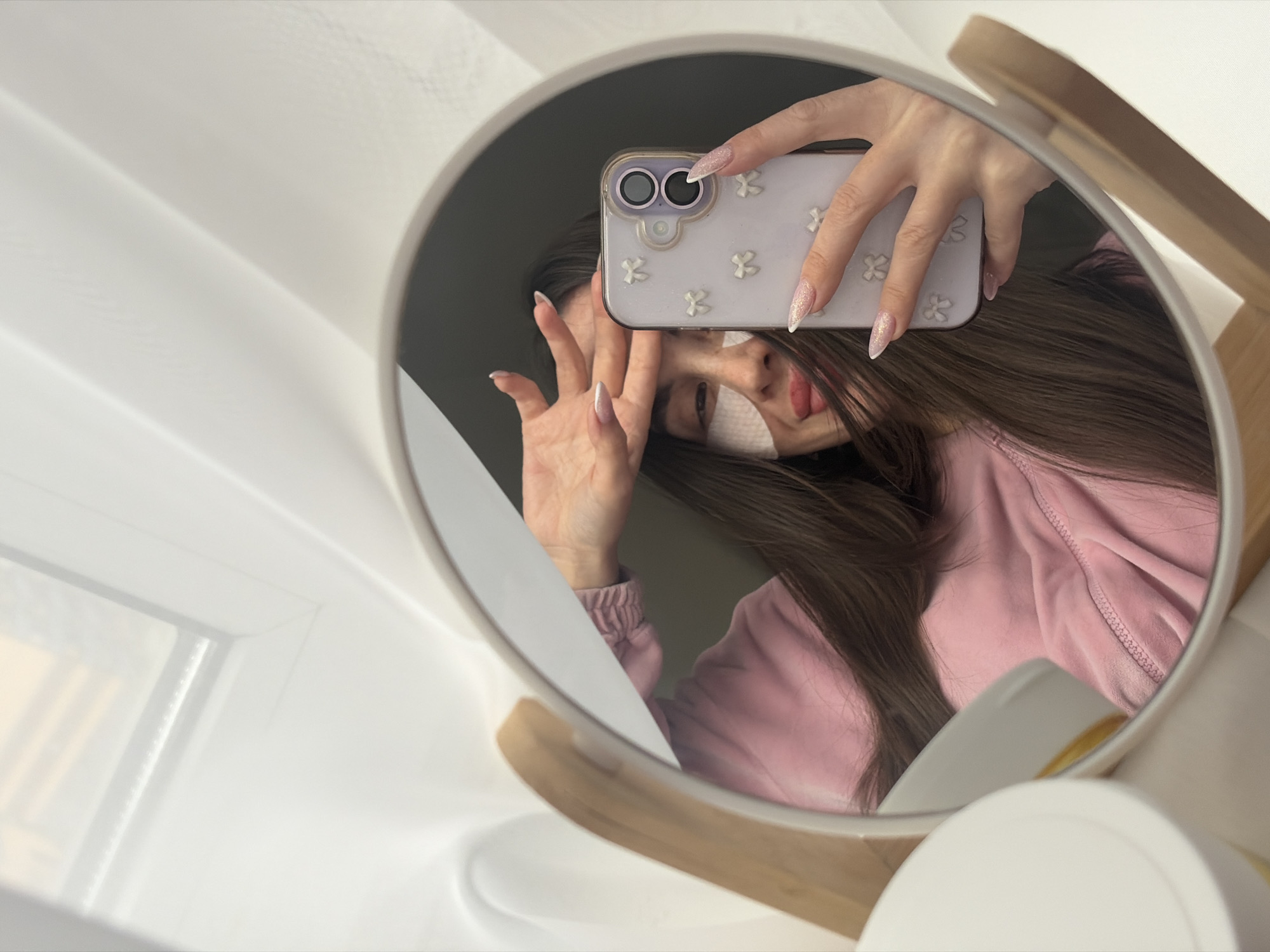 The image is a selfie taken in a mirror, showing a person from the chest up. The person is wearing a pink, velour-like sweater and has long, brown hair. Adhesive strips are visible under their right eye. They are holding a white smartphone with a floral-patterned case in front of their face. The nails on the hand holding the phone are long and pointed, with a glittery finish. The mirror is round and supported by a wooden stand. The background includes a white windowsill and parts of a white table or counter. The image does not provide enough context to determine a specific location in Saint Petersburg, Russia.
