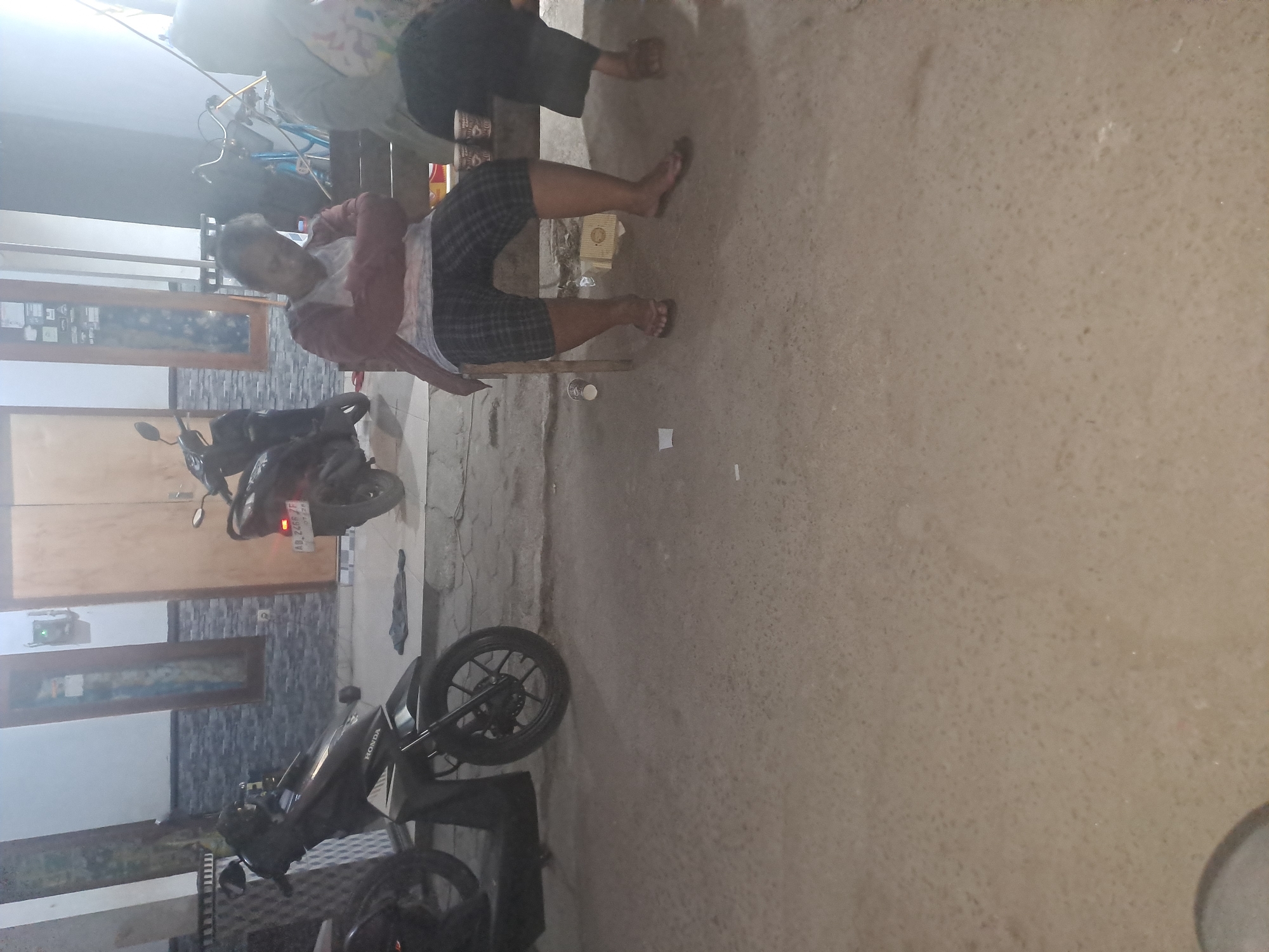 The image depicts an outdoor scene, likely in Tirtomartani, Indonesia, at dusk or night. In the foreground, a concrete or asphalt surface is visible. To the left, the rear of a black motorcycle with a prominent "HONDA" logo is parked. In the middle ground, a wooden bench is occupied by two individuals. The person on the left, wearing a red and white long-sleeved shirt and checkered shorts, is seated with their arms crossed and head bowed, appearing to be resting or sleeping. The person on the right is mostly obscured, wearing a light-colored top and dark skirt, with their legs visible as they sit. Various objects are scattered around the bench, including paper cups and a small yellow box. In the background, a building with large doors and windows is present. Another motorcycle is parked near the building, with a license plate visible that reads "AD 2466 Zf". A bicycle is also partially visible in the background. The overall lighting suggests it is evening.