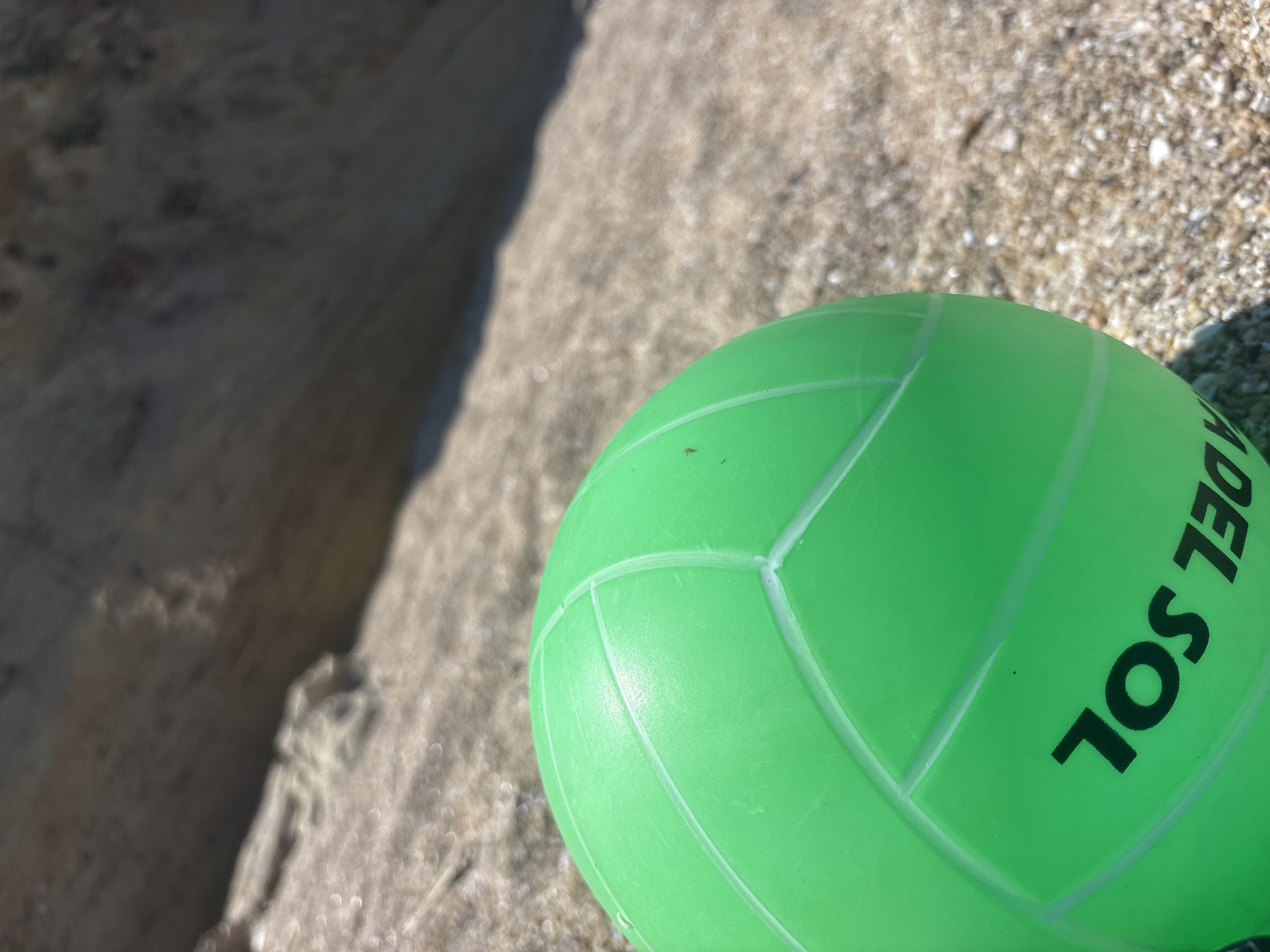 A bright green volleyball rests on a sandy beach next to a rock formation. The volleyball is the primary subject, and its surface displays white lines and the black text "DEL SOL". The sand has a rough texture, with small shells and pebbles visible. The rock formation appears to be a light, rough-textured stone. The lighting suggests it is daytime, likely outdoors. The scene is set on a beach, with the implied presence of the sea or ocean nearby given the sand and rock. No people or animals are visible in the frame. The image is a close-up shot, focusing on the volleyball and the immediate surroundings, with the background blurred. The overall mood is calm and relaxed, characteristic of a beach setting. The location is Burgas, Bulgaria.