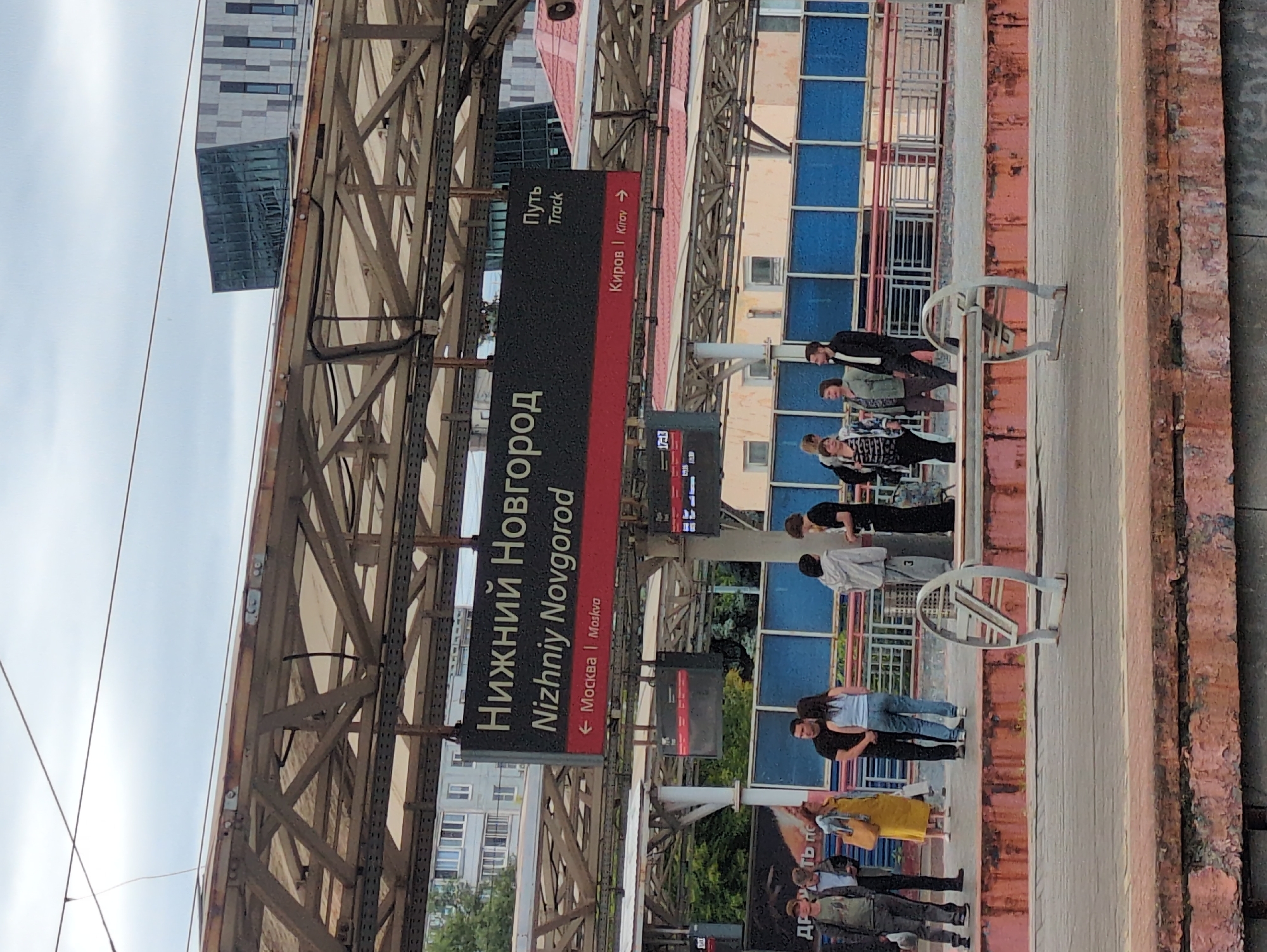 This media file depicts a platform at a train station in Nizhny Novgorod, Russia. The scene is viewed from an angle, with a large sign prominently displaying "Нижний Новгород" (Nizhny Novgorod) and its English translation, "Nizhniy Novgorod". Other text on signs indicates "Путь Track" and directions to "Москва | Moskva" (Moscow) and "Киров | Kirov".
Several people are visible on the platform, some standing in small groups, others walking. They appear to be waiting for a train or disembarking. The overall atmosphere suggests a typical day at a busy transportation hub. The sky is overcast, indicating a cloudy day. The architecture of the station is visible, featuring metal structures and a building with blue-tinted windows. The presence of train tracks and overhead wires confirms the setting is a functional train station. One sign shows a time of "17:13", suggesting it is late afternoon. There are no strong emotional expressions visible on the faces of the people, their demeanor appearing neutral and focused on their travel.