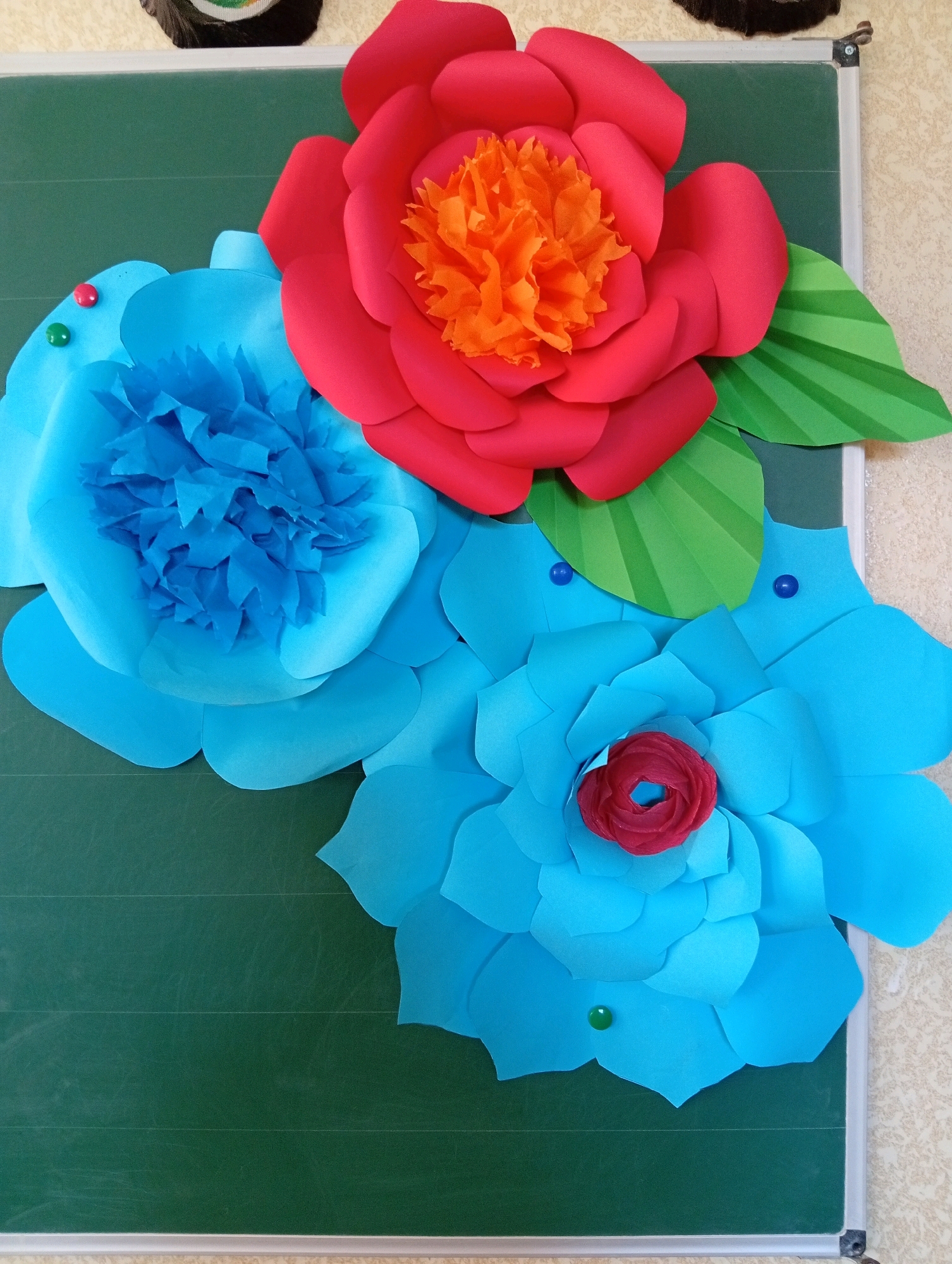 An arrangement of large, colorful paper flowers is affixed to a green chalkboard. The flowers are in shades of red, blue, and green. The red flower is the largest, with bright orange tissue paper in its center. There are two large blue flowers, one with a lighter blue, crinkled center and the other with a smaller red flower in its center. Green paper leaves are also part of the arrangement. Several small, colored pushpins are used to attach the flowers to the board. The chalkboard has faint horizontal lines and is framed by a silver metal edge. The background wall appears to be a textured beige wallpaper. The image is taken from a slightly elevated, head-on perspective.