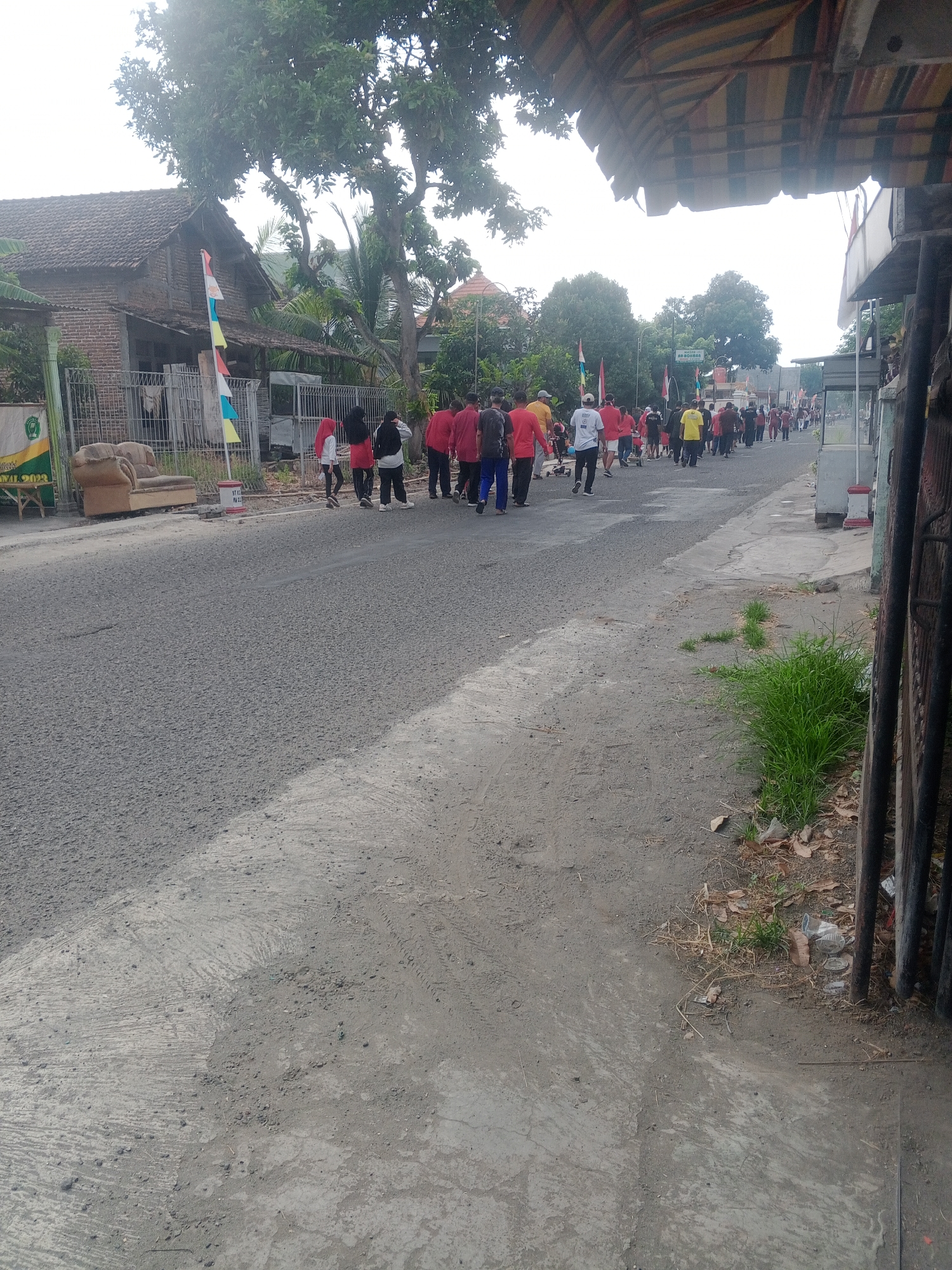 In Lembah, Indonesia, a group of people walk down a paved road, participating in a procession. The road is lined with buildings and vegetation, with a large tree casting shade over a portion of the scene. The people are dressed in casual attire, with some individuals wearing red shirts. They are walking in the same direction, suggesting a coordinated event. The environment appears to be a residential or semi-urban area, with a mixture of houses, fences, and open spaces. The overall atmosphere is one of community gathering or a local event.