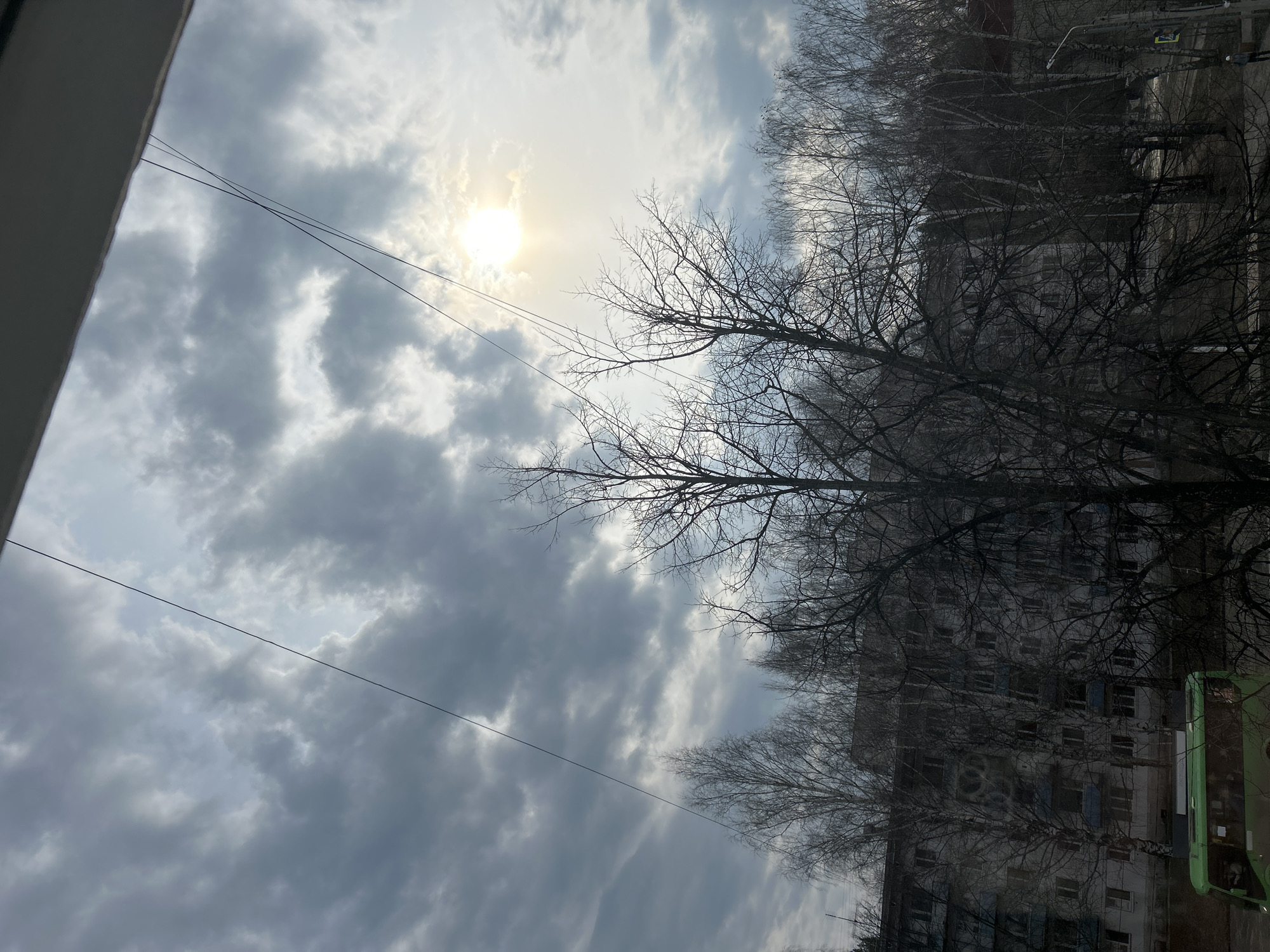 The image, taken in what appears to be the city of Chaikovsky, Russia, shows a view from a window. The sky is predominantly gray with overcast clouds, but the sun is visible behind the clouds. In the foreground, there are power lines. Behind the power lines, there are bare trees without leaves. Beyond the trees is a multi-story building. A green vehicle is also visible to the right of the building. The scene appears to be taken during the late fall or winter season.
