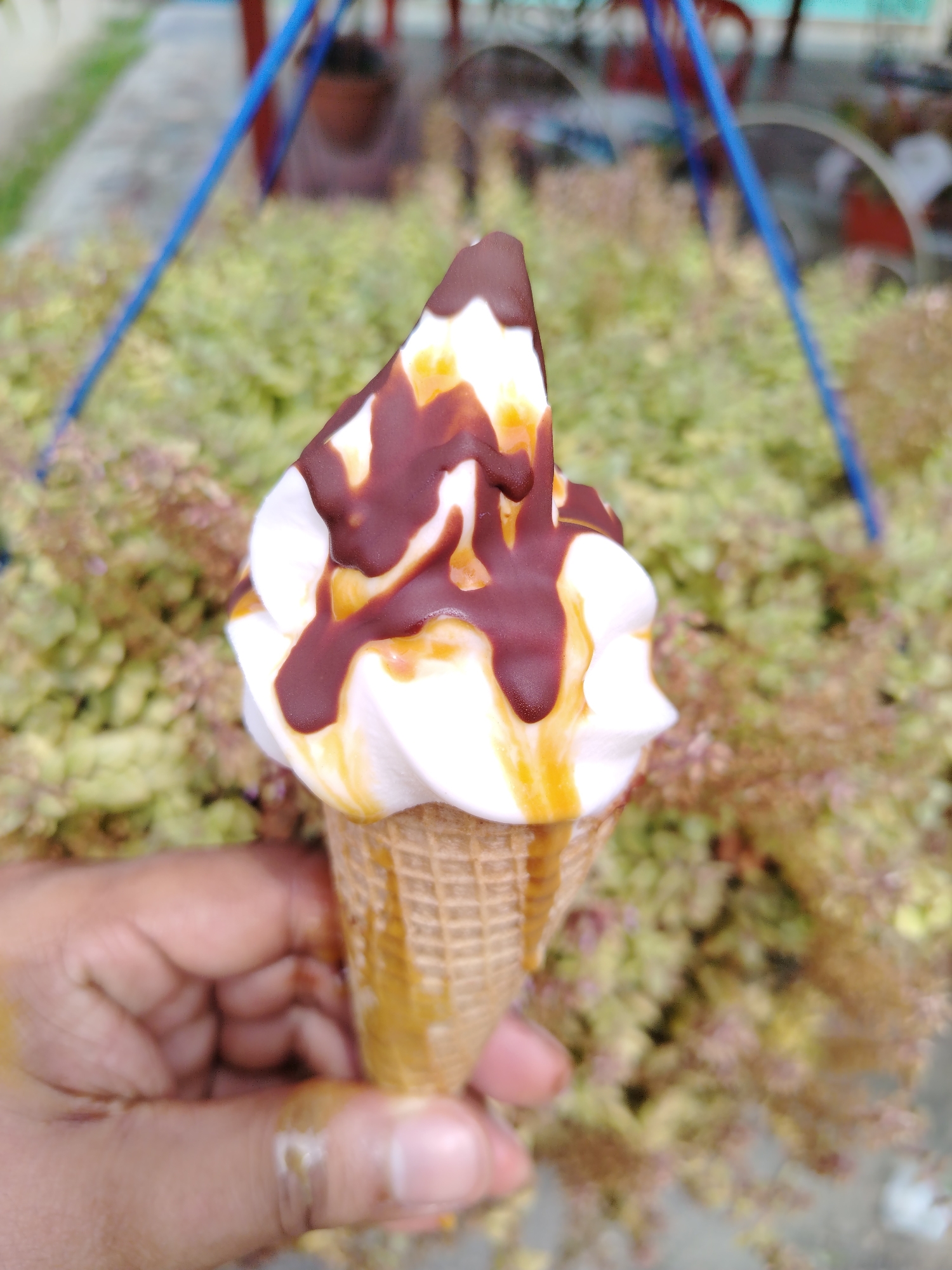 A person holds an ice cream cone with soft-serve ice cream drizzled with chocolate and caramel. The ice cream is in front of a leafy green and brown bush, with a blue frame structure visible in the background. The image was taken in Tinaquillo, Venezuela.