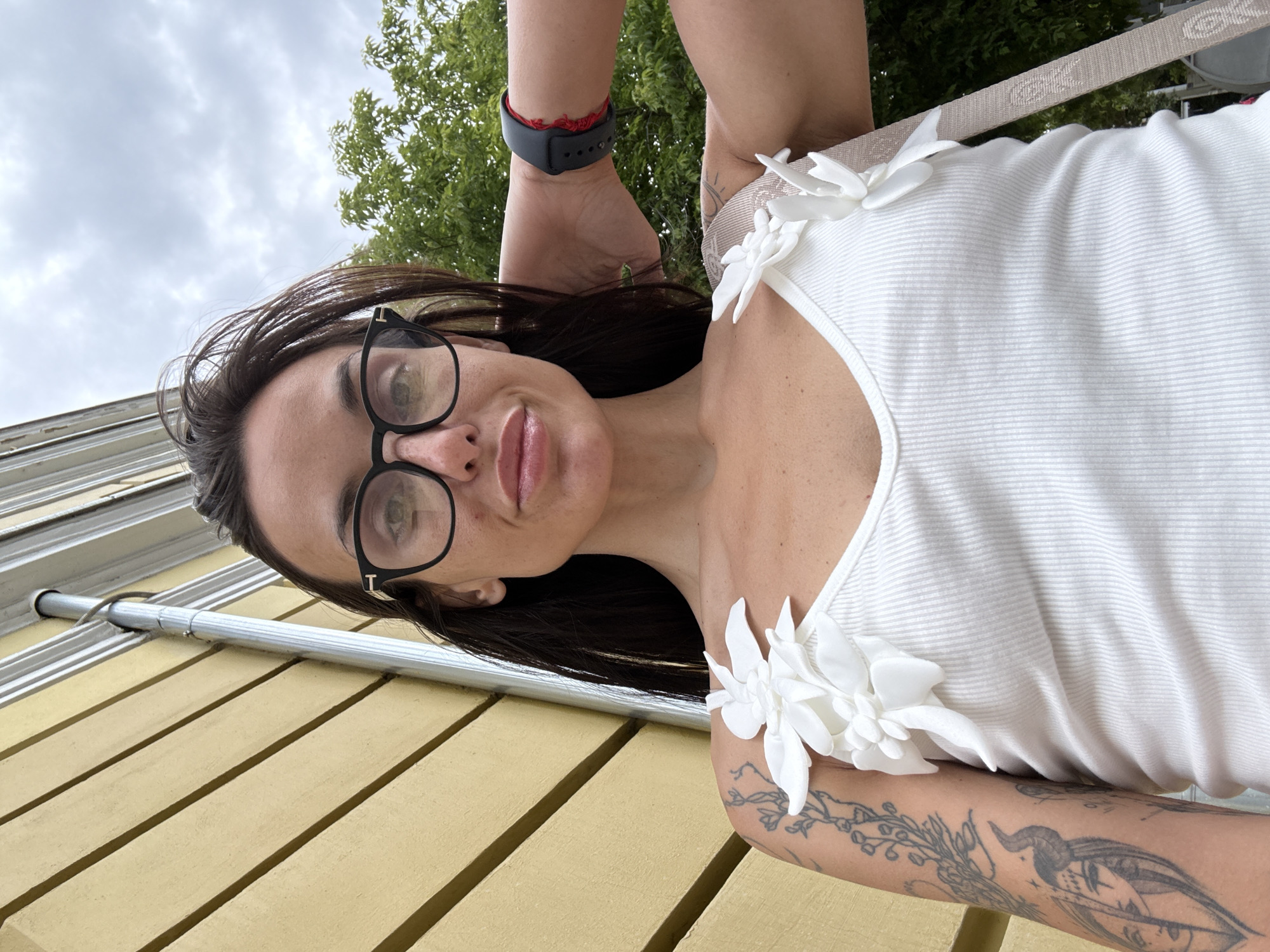 A woman with long, dark hair and glasses is standing outdoors, facing the camera. She is wearing a white tank top adorned with white floral appliques on the straps. She has tattoos on her left arm. A smartwatch is visible on her right wrist. Behind her, a yellow building with horizontal siding is partially visible, along with a metal downspout. Green trees and a cloudy sky form the background. The scene appears to be on a street in Pleven, Bulgaria.