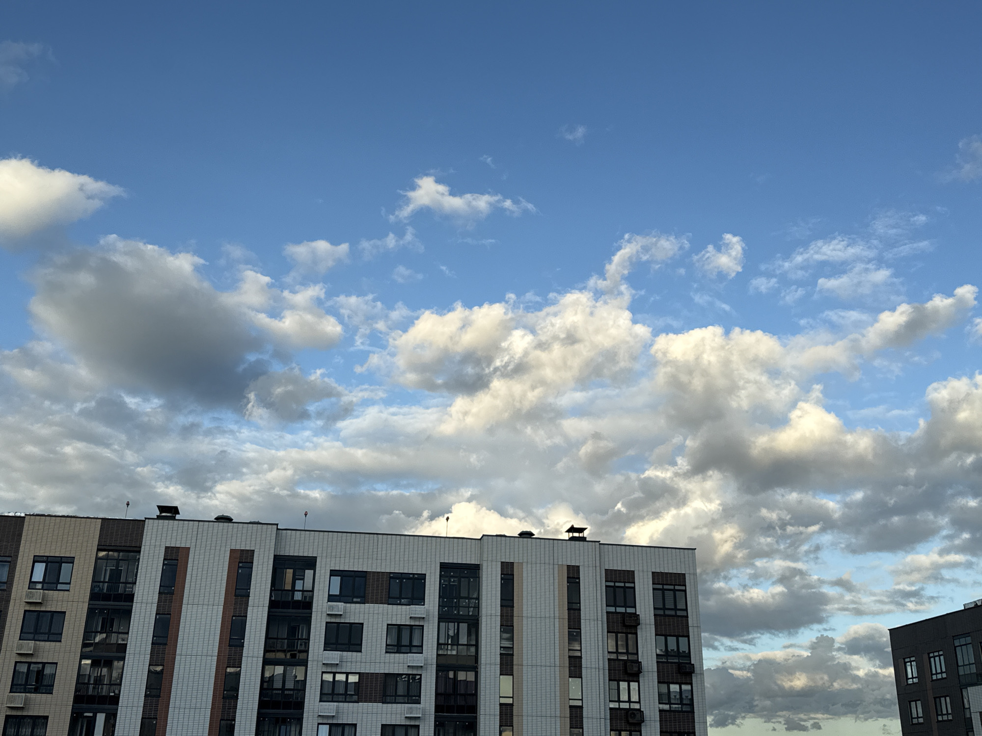 The image captures a scene in Lyubertsy, Russia, featuring a modern apartment building against a backdrop of a dynamic sky. The building, which appears to be a multi-story residential complex, is characterized by its facade adorned with a mix of white, gray, and brick-colored tiles. Numerous dark-framed windows and outdoor air conditioning units are visible across its several floors, suggesting it is occupied. The roofline features ventilation shafts and what appear to be small red warning lights. To the right, a portion of another, darker building is visible. Above the structures, the sky is a vibrant blue, populated by a variety of clouds ranging from bright white, sunlit formations to darker, more diffused gray clouds. The lighting suggests it is either late afternoon or early evening, with the sun illuminating the clouds from an angle. There are no people or discernible actions taking place in the image. The scene is static, focusing on the architecture and the atmospheric conditions.