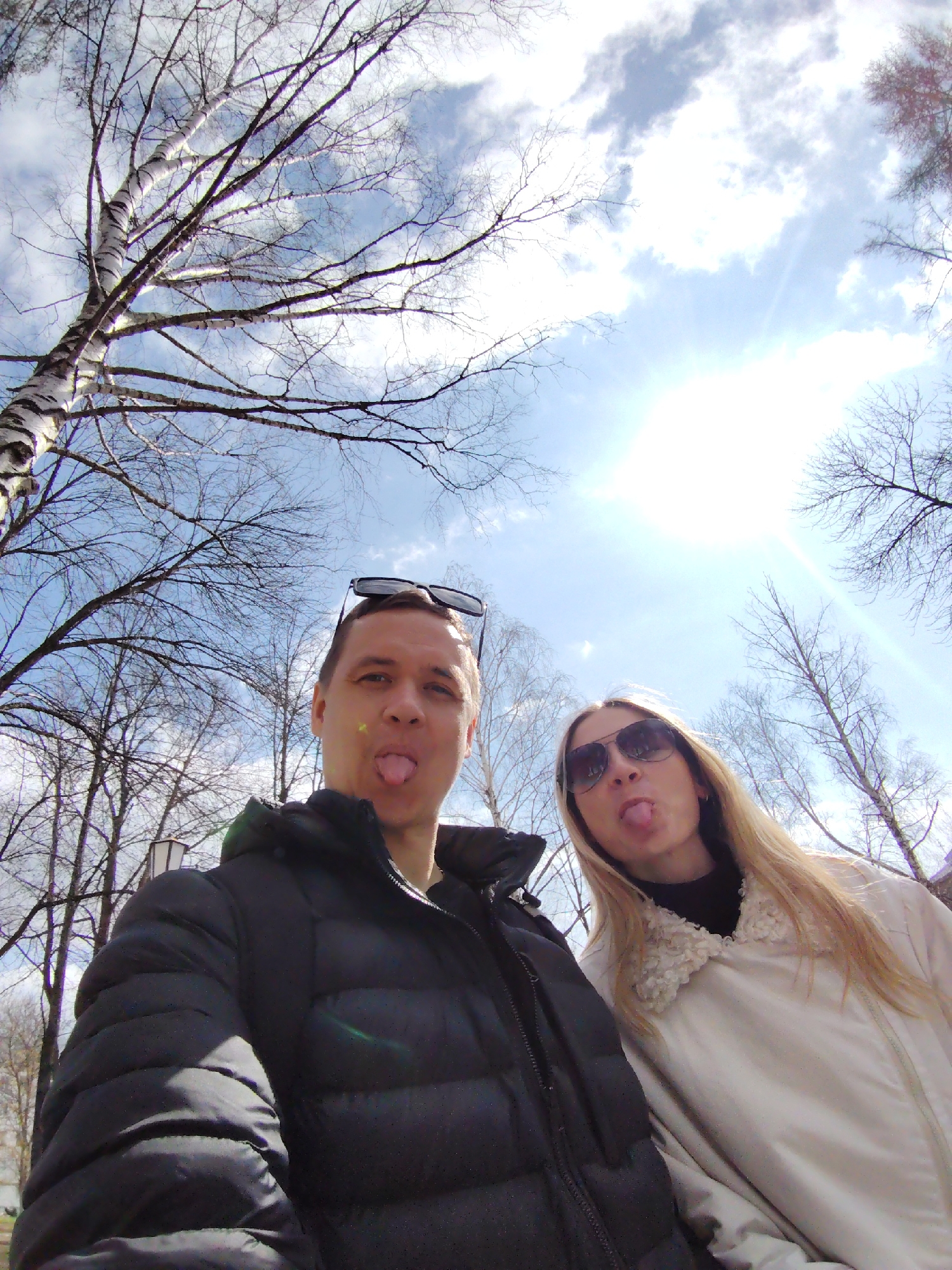 A low-angle selfie taken in Chaikovsky, Russia, captures two individuals sticking out their tongues. The man on the left wears a black puffy jacket and has sunglasses resting on his head. The woman to his right sports a white jacket with a textured collar and dark sunglasses. The backdrop includes leafless trees and a bright, partly cloudy sky with the sun shining prominently.