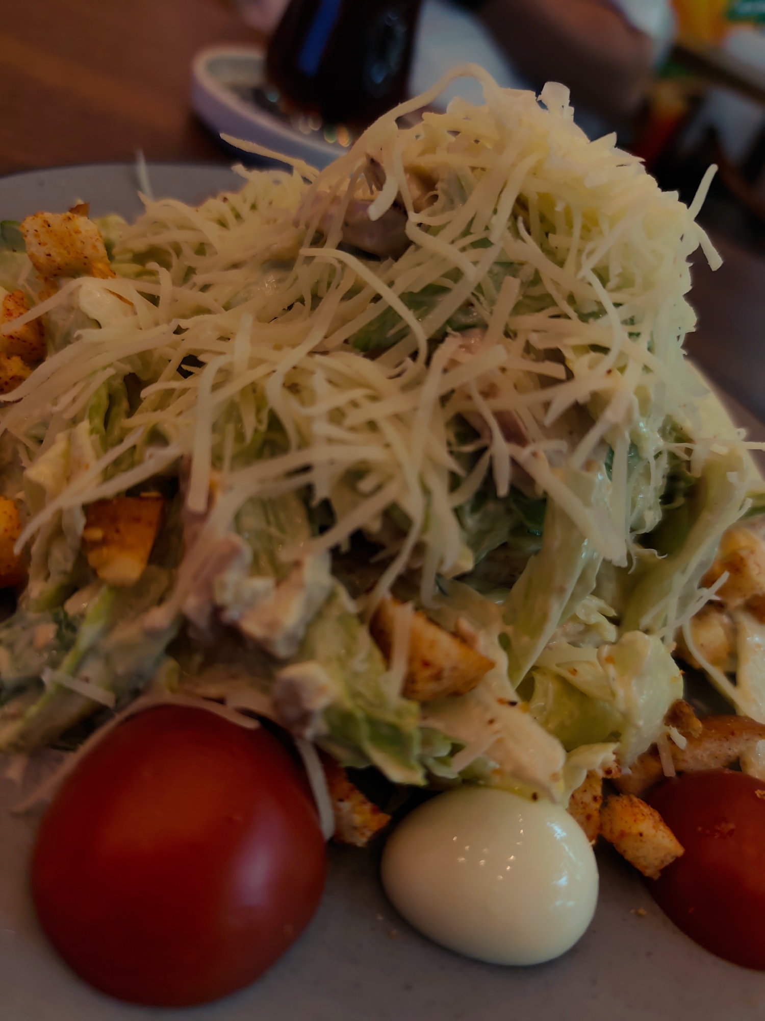 A close-up shot shows a plate of Caesar salad topped with shredded cheese. The salad appears to contain lettuce, croutons, and possibly chicken. In the foreground, there are two whole cherry tomatoes and one peeled hard-boiled egg. The salad is served on a gray plate, which rests on a wooden table. The background is blurred, showing hints of other dishes and a dark bottle. The lighting is dim, creating a warm and intimate atmosphere. There are no people visible in the image. The image does not provide information about the location in Fergana City, Uzbekistan.