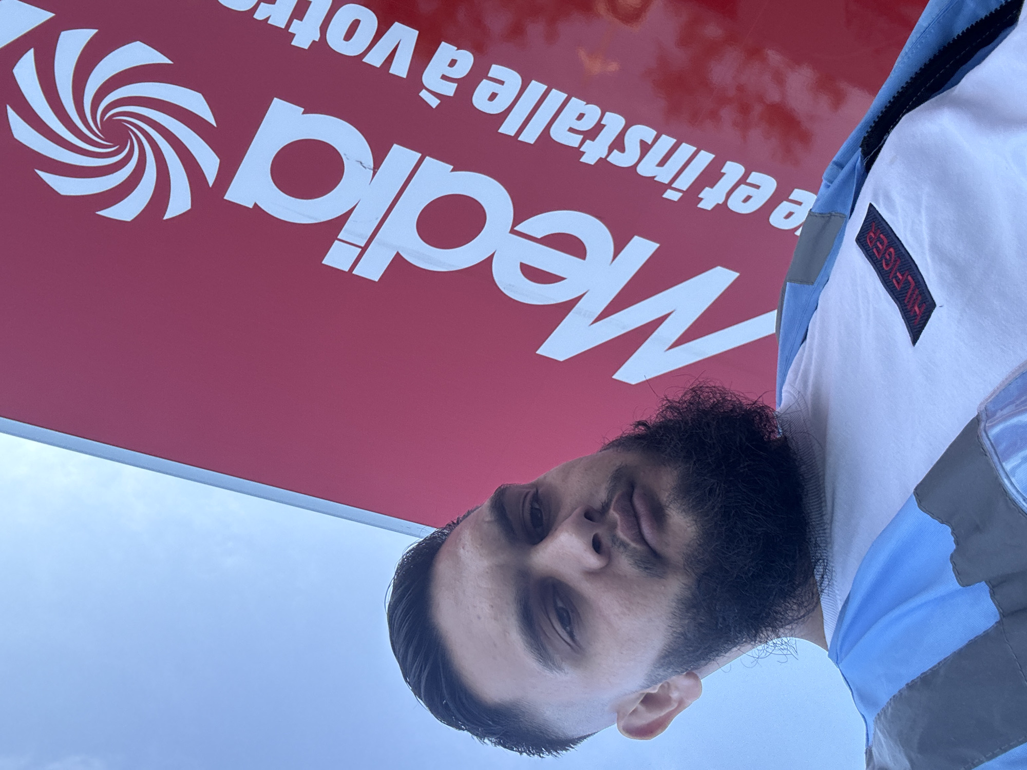 A man with dark hair, a beard, and a fair complexion is captured in a low-angle, selfie-style photograph. He is wearing a white t-shirt with a rectangular red and black patch that reads "HILFIGER" on the left chest area, and a blue and grey jacket. The man is positioned in front of a large, vibrant red sign with white text and a swirling white logo. The sign appears to be advertising a company named "Media" and also includes the text "e et installe à votre". The sky is visible above the man and the sign, appearing light blue with some faint cloud formations. The overall lighting suggests it is daytime. The location is specified as the city of Boom, Belgium.