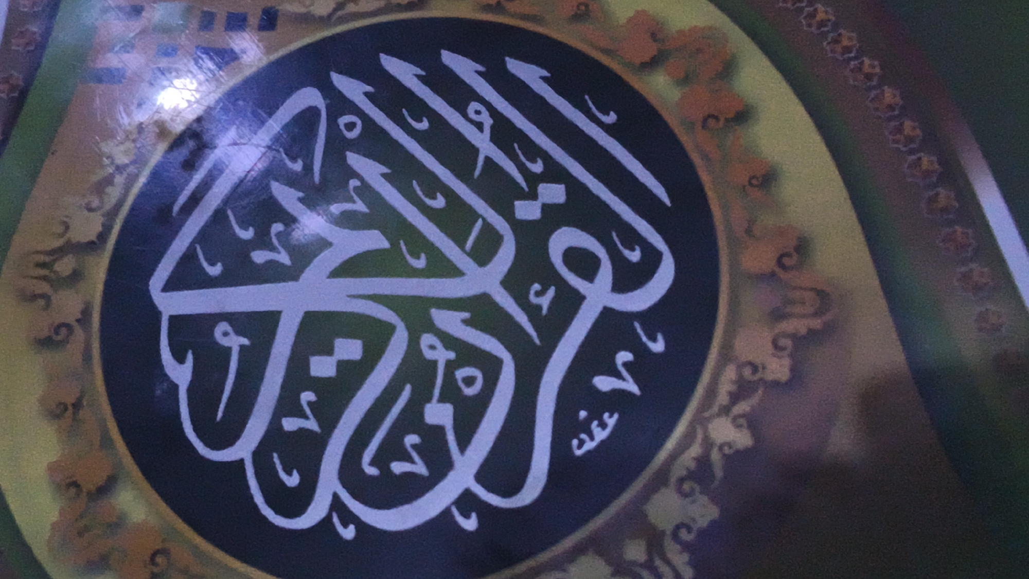 The image shows a close-up of Islamic calligraphy, likely from the Quran, rendered in white against a dark circular background. The calligraphy is ornate and stylized, with flowing lines and intricate letterforms. Surrounding the central circle is a decorative border featuring floral or paisley-like motifs in shades of orange and yellow. The overall impression is one of religious art and ornamentation, possibly part of a larger decoration or object. The image appears to be taken in low light, with some glare on the surface. The location of Kerep Kidul, Indonesia, is not directly evident from the visual content of the image itself, but it is provided as contextual information.