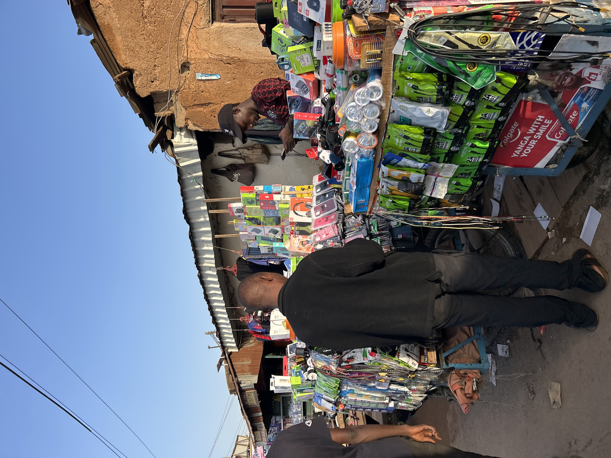 This outdoor scene, likely in a market area of Jos, Nigeria, captures a street-level view of a mobile phone accessories vendor. The vendor, wearing a red and black patterned shirt and a black baseball cap, is occupied with his phone while standing behind a pushcart overflowing with merchandise. The cart showcases a variety of mobile accessories, including chargers, cables, phone cases, and headphones, neatly arranged. A 'Colgate' advertisement with the slogan "YANGA WITH YOUR SMILE" is visible on the side of the cart. An African man in a black shirt is standing, examining the merchandise on the cart. The scene is framed by weathered buildings on either side and a clear blue sky above, indicating daytime. The ground is paved and dusty.