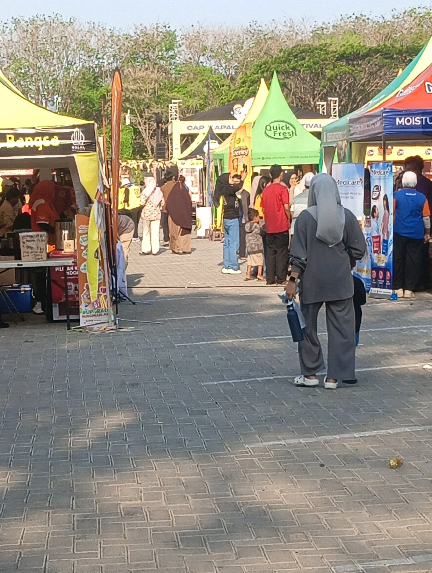 This is a medium shot of an outdoor market or festival scene. In the foreground, two individuals are walking away from the camera, one in a grey hijab and matching outfit, carrying a dark water bottle. To the left, a child in a grey and white outfit is being held by an adult, also facing away. Behind them, a crowd of people browses various stalls, characterized by colorful tents. One tent on the left displays "Bangsa" and "HALAL" logos. Another tent, predominantly green, features a "Quick Fresh" logo. Further back, a blue tent displays "MOISTU" and "Medicare" branding. The ground is paved with interlocking grey bricks, and the background shows trees and what appears to be event structures. The presence of "HALAL" and the general ambiance suggests a cultural or religious festival, possibly related to Islamic traditions. The text "Surabaya, Indonesia" is not directly visible in the image, but the context of a bustling market with food and consumer goods aligns with typical Indonesian street or festival scenes.