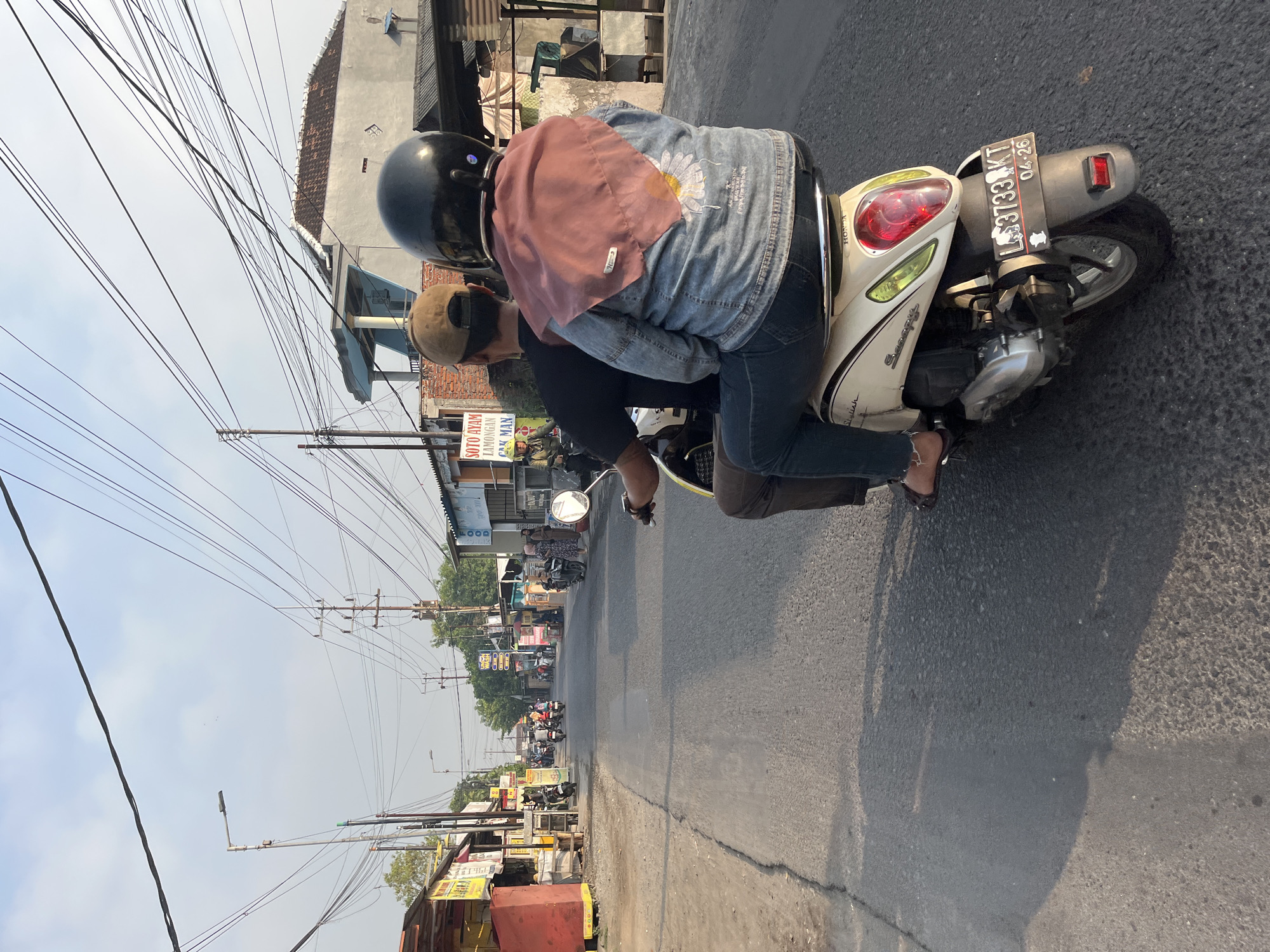 In Kedungkendo, Indonesia, two individuals are riding a light-colored Honda Scoopy motorcycle along a paved road. The rider, wearing a brown cap and a black t-shirt, is in the front. The passenger behind, wearing a pink hijab, a denim jacket with a floral design, and jeans, is seated at the back. The motorcycle has a license plate that reads "L 3733 KT 04-26". The street is lined with small businesses and residential buildings. Overhead, a dense network of power lines crisscrosses the sky. The sun casts shadows on the road, suggesting it is either early morning or late afternoon. The overall atmosphere is that of a typical day in a bustling Indonesian town.