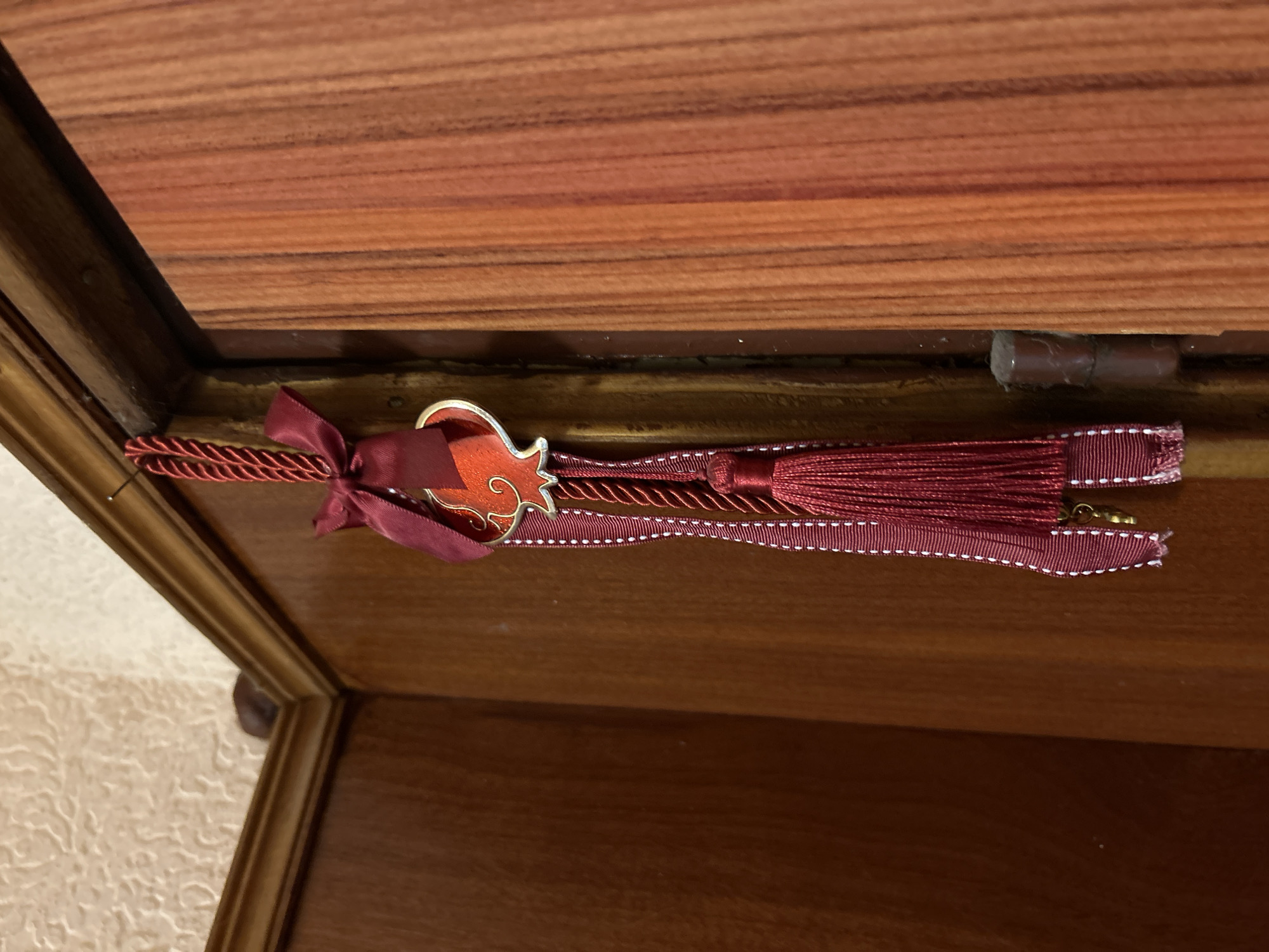 A close-up shot depicts a burgundy-colored ornament attached to the interior of a wooden box. The ornament features a stylized fish-shaped charm in orange with a gold outline, suspended from a burgundy ribbon tied into a small bow. It is attached to a burgundy tassel that is attached to a burgundy ribbon with a white, dashed line detail. The box's interior shows a wood grain pattern with shades of brown and red. A hinge for the box's lid is visible on the right side. The box's exterior has a glossy golden brown finish and stands on a surface with wallpaper. There is no indication of the image's location.