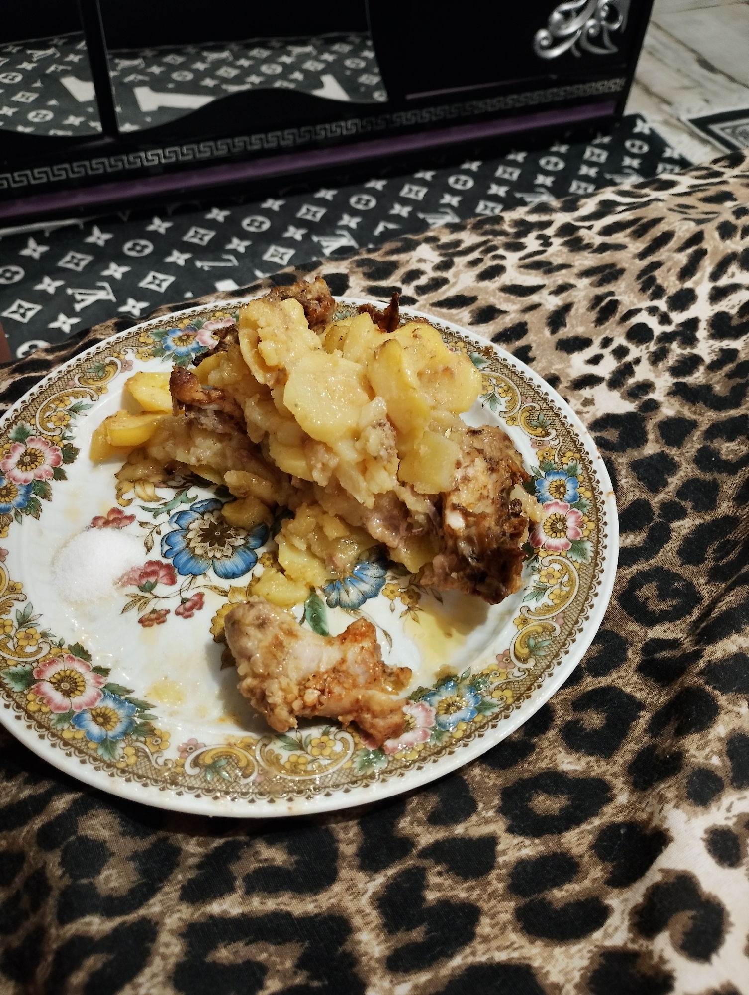A close-up shot shows a plate of food, likely chicken and potatoes, on a table covered with a leopard-print cloth. A pile of the food is in the center of the plate, with a small amount of salt to the left. The plate has a floral pattern around the rim. In the background, a dark, ornate object with reflective surfaces is visible. The overall impression is of a home-cooked meal served casually. The city of Zimnitsa, Bulgaria is not depicted in the image.