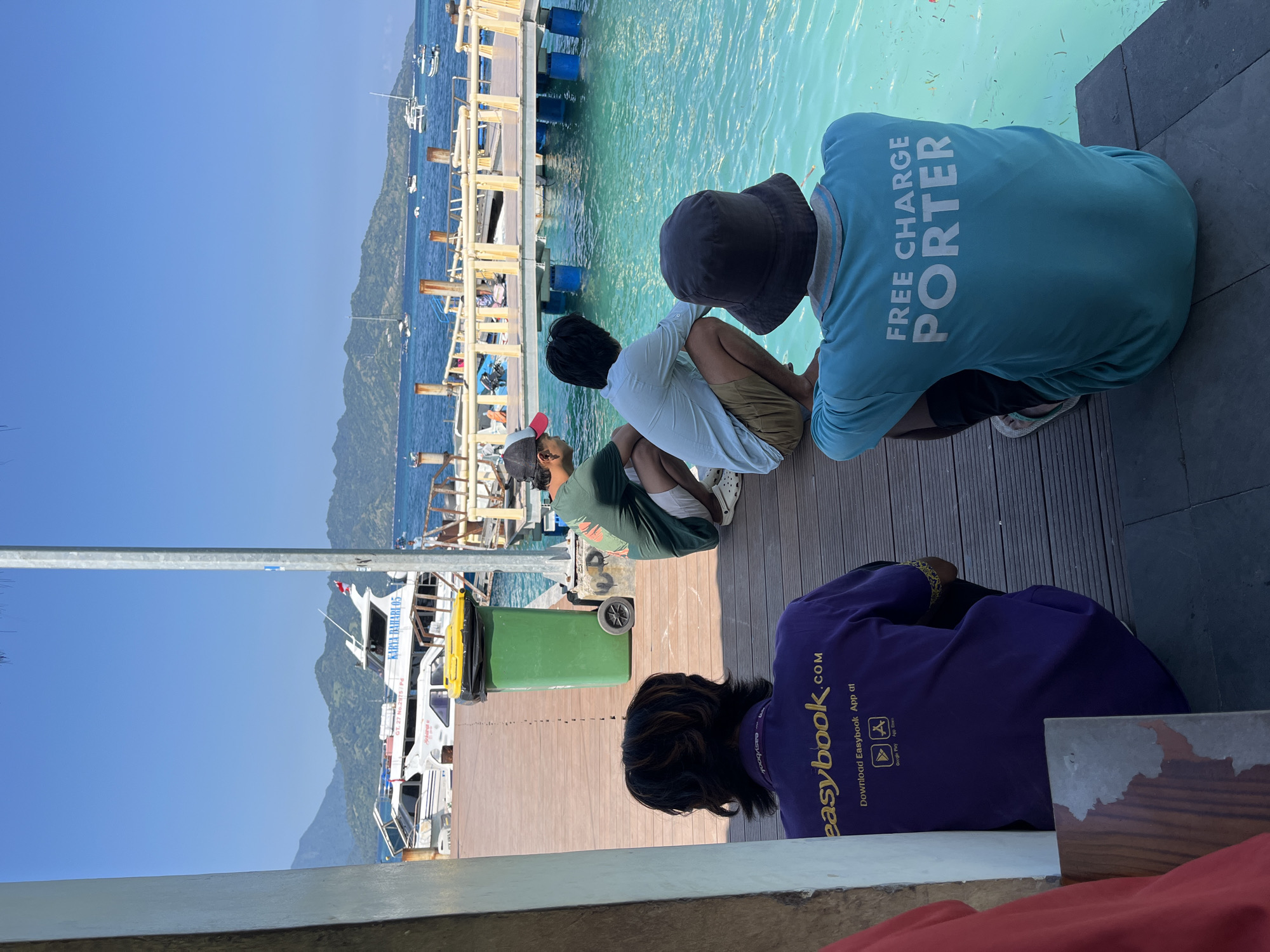 The photograph depicts a waterside scene in an unidentified location of Unknown, Indonesia. Four individuals are observed sitting on a wooden deck or dock. One person wears a light blue shirt with "FREE CHARGE PORTER" printed on the back, another has on a purple shirt displaying "easybook.com," and two other individuals are situated between them. A green trash can sits on the deck. In the background, docked boats can be seen, along with a mountain range. The sea exhibits a clear, turquoise hue. The sky is bright blue, suggesting a clear day.