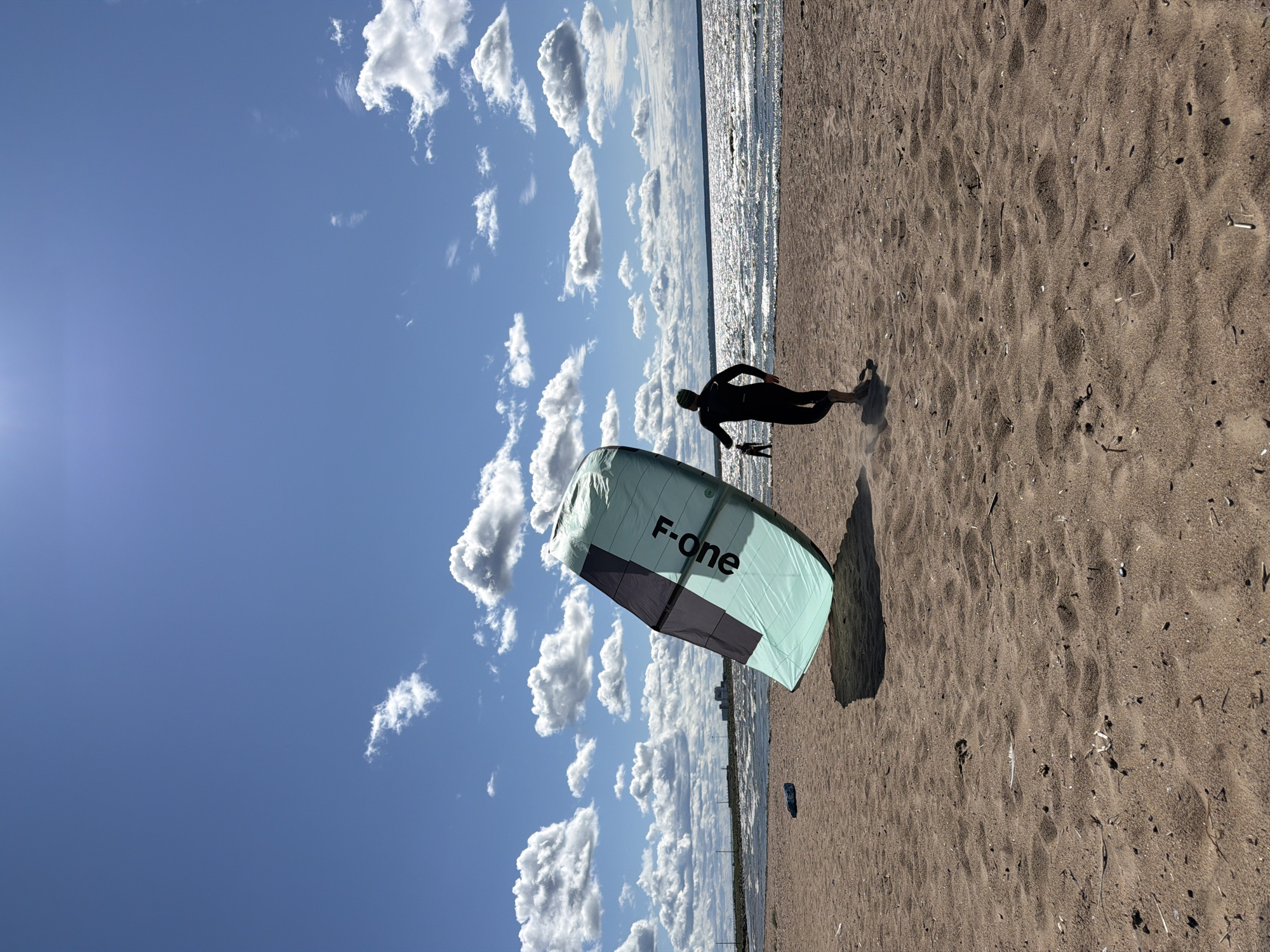 A person in a black wetsuit stands on a sandy beach next to a large, mint green and black kite. The kite is angled, with the text "F-one" visible in black lettering. The person is facing away from the camera, moving towards the water. The beach is textured with sand and scattered debris. In the background, a vast body of water shimmers under a bright blue sky filled with scattered white clouds. A distant shoreline is visible, with a building structure on the horizon. The scene appears to be a beach in Kronstadt, Russia, on a sunny day suitable for watersports.