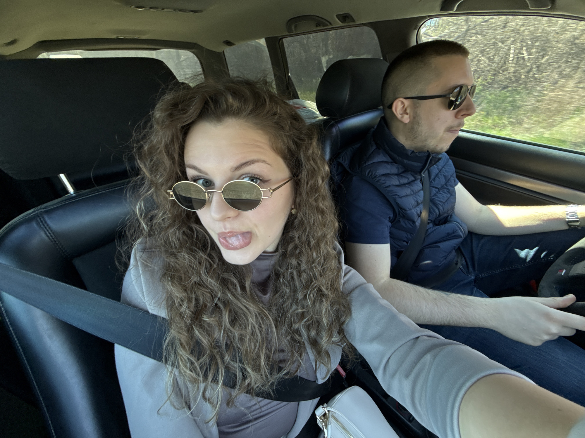 A man and a woman are in the front seats of a car. The woman is in the passenger seat wearing sunglasses, a gray top, and a seatbelt. She is sticking her tongue out. The man is driving, wearing sunglasses, a navy blue vest, a dark blue shirt, blue jeans, and a wristwatch. The location of the image is not confirmed to be in the city of Nikolaevo, Bulgaria.