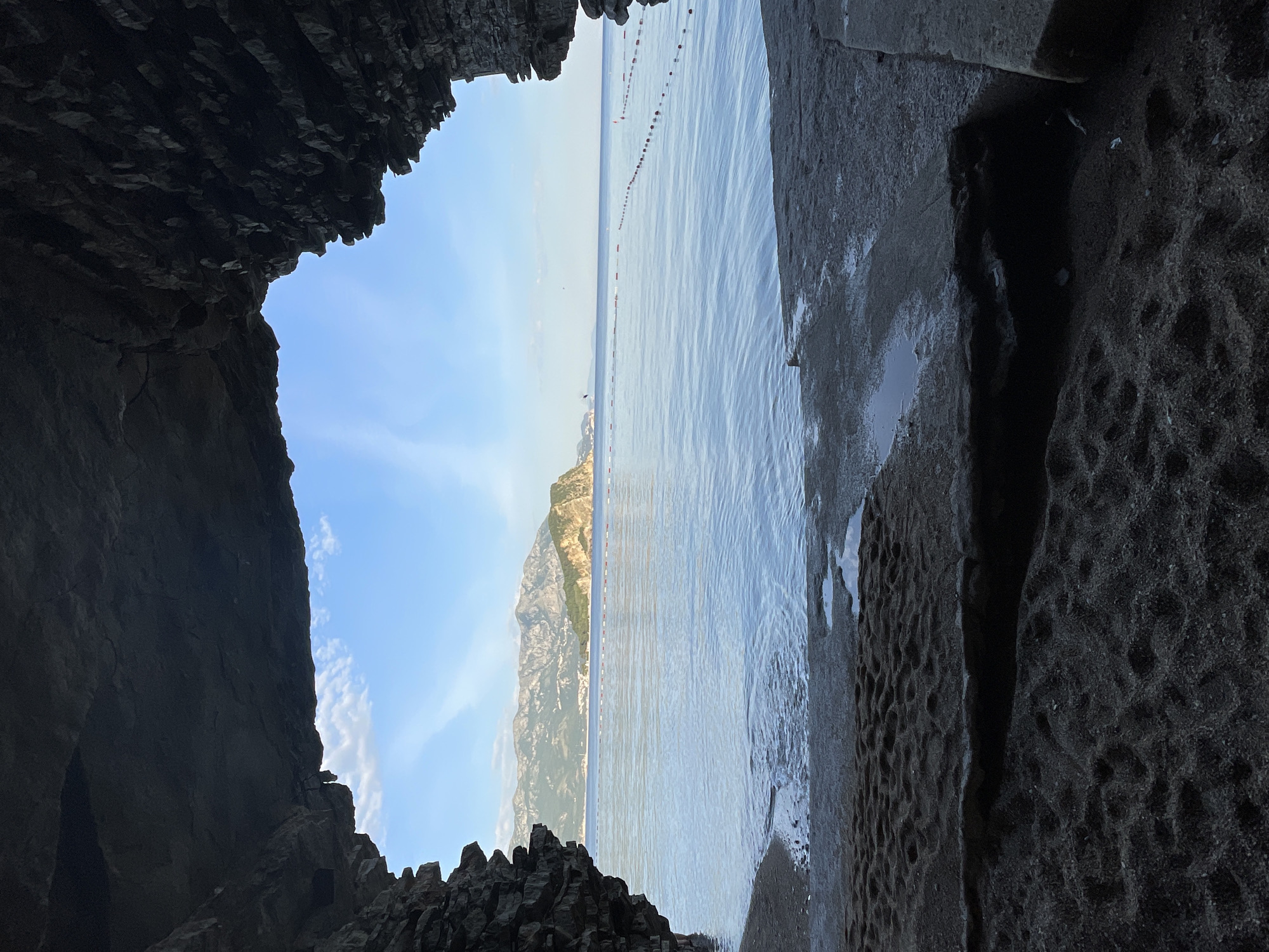 View from inside a dark cave in Sveti Stefan, Montenegro, looking out to the Adriatic Sea. A line of red and white buoys stretches across the water. In the distance is a mountain range. The sky is blue with scattered clouds. The cave entrance is jagged and uneven.