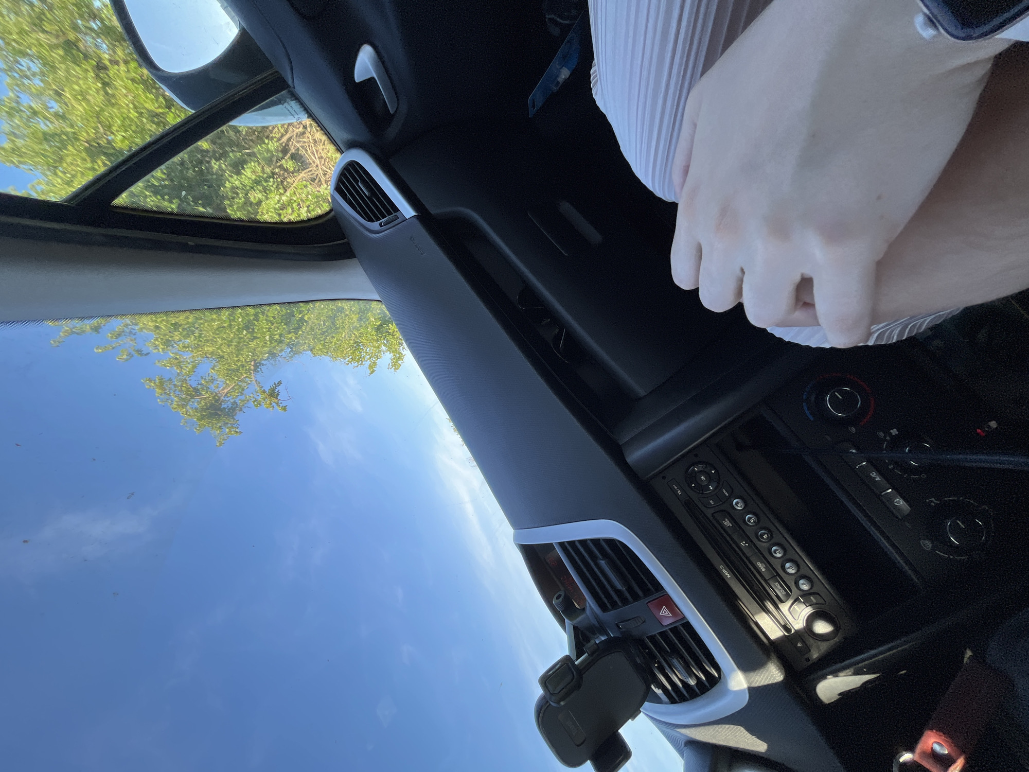 The image shows the interior of a car, specifically the dashboard and a person's hands. The dashboard is black with silver accents and includes air vents, a phone mount, and the car's radio and climate control panel. A pair of pale hands is visible, wearing what appears to be a white and gray striped shirt or blouse. The sky is visible through the windshield, with a few trees visible, too. The location within the city of Unknown, Unknown is not identifiable.