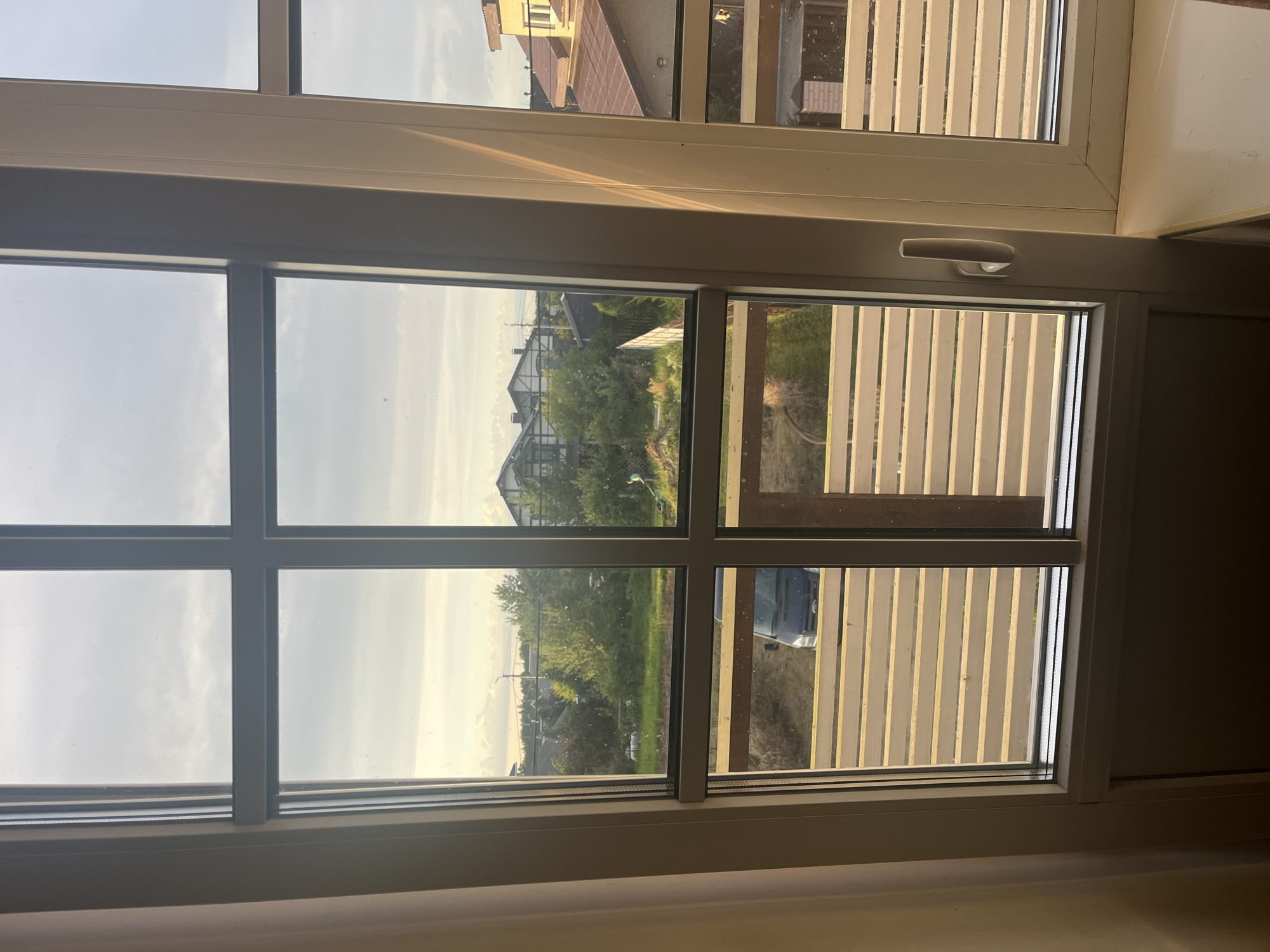 The image depicts a view through a multi-paned window, looking out onto a suburban residential area. The window frames are white, and the glass is clean, allowing for a clear view of the exterior. In the foreground, the lower portion of the window reveals a light-colored, slatted wooden structure, possibly a balcony or fence.
Beyond this, the scene opens up to a green yard with scattered trees and several houses with pitched roofs, typical of residential architecture. Some of the houses are multi-story, with visible windows and architectural details. One house in the mid-ground has a distinctive white and dark trim design.
In the yard, a dark-colored van is parked, and to the right, a person can be seen in the distance, appearing to be engaged in outdoor activity, possibly mowing grass. The sky is partly cloudy with a soft, diffused light, suggesting it might be late afternoon or early morning. The overall impression is of a peaceful, everyday scene in a residential setting.
The location is identified as Vladimirovka, Russia.