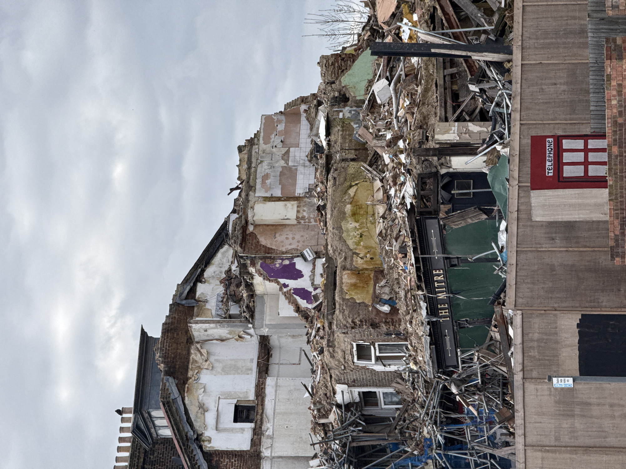The image shows a demolished building in London, United Kingdom. The structure is a mix of brick and concrete, and the interior walls are visible. The signage "THE MITRE" suggests the building was a pub or bar. A red painted telephone box is seen. The scene is surrounded by debris and scaffolding, indicating ongoing demolition work. The sky is overcast, implying a somber atmosphere. No people are visible in the frame.