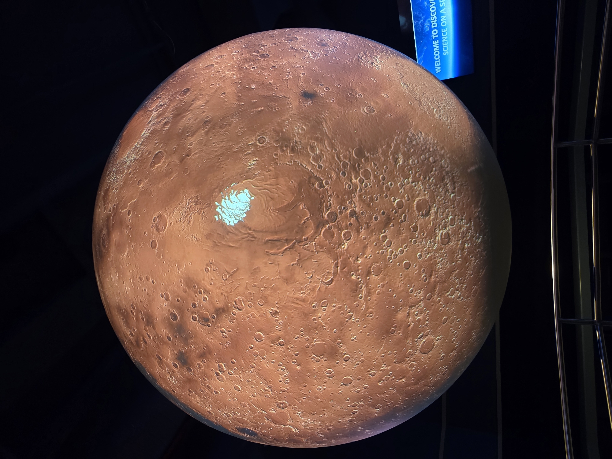 The image shows a large, spherical projection of the planet Mars, suspended in a dark environment, likely an exhibit or planetarium. The surface of Mars is detailed with craters, mountainous regions, and what appears to be a polar ice cap, rendered in shades of orange, brown, and a bright blueish-white. In the lower right corner, a blue sign with white text is visible, which reads "WELCOME TO DISCOVE SCIENCE ON A SE". This suggests the location is likely an educational institution or science center, possibly related to astronomy or space exploration. The perspective is from below, looking up at the planet. Metal railings are visible in the foreground, indicating a viewing area for visitors. The overall impression is one of an immersive educational display. The location is identified as Santa Ana, United States of America.