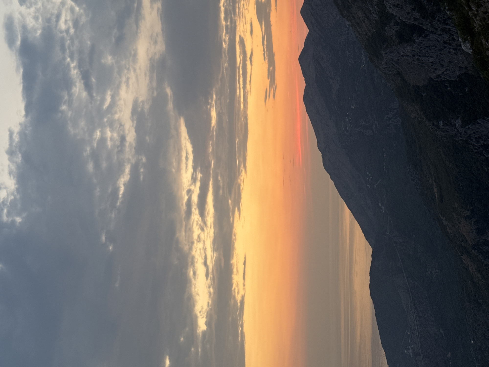The image depicts a dramatic mountain landscape at sunset, possibly near Gluhi Do, Montenegro. The sky is a gradient of colors, transitioning from a pale yellow and orange glow near the horizon to dark, cloudy gray at the top. Wispy clouds are scattered across the sky. Below the sky, a body of water, likely the sea, reflects the warm hues of the sunset. Dominating the foreground are dark, silhouetted mountains, their rugged peaks and slopes sharply defined against the brighter sky. The mountains appear to be sparsely populated, with a few faint lights visible in the distance, suggesting small settlements or scattered houses on the slopes. The overall impression is one of serene, natural beauty at the end of the day.